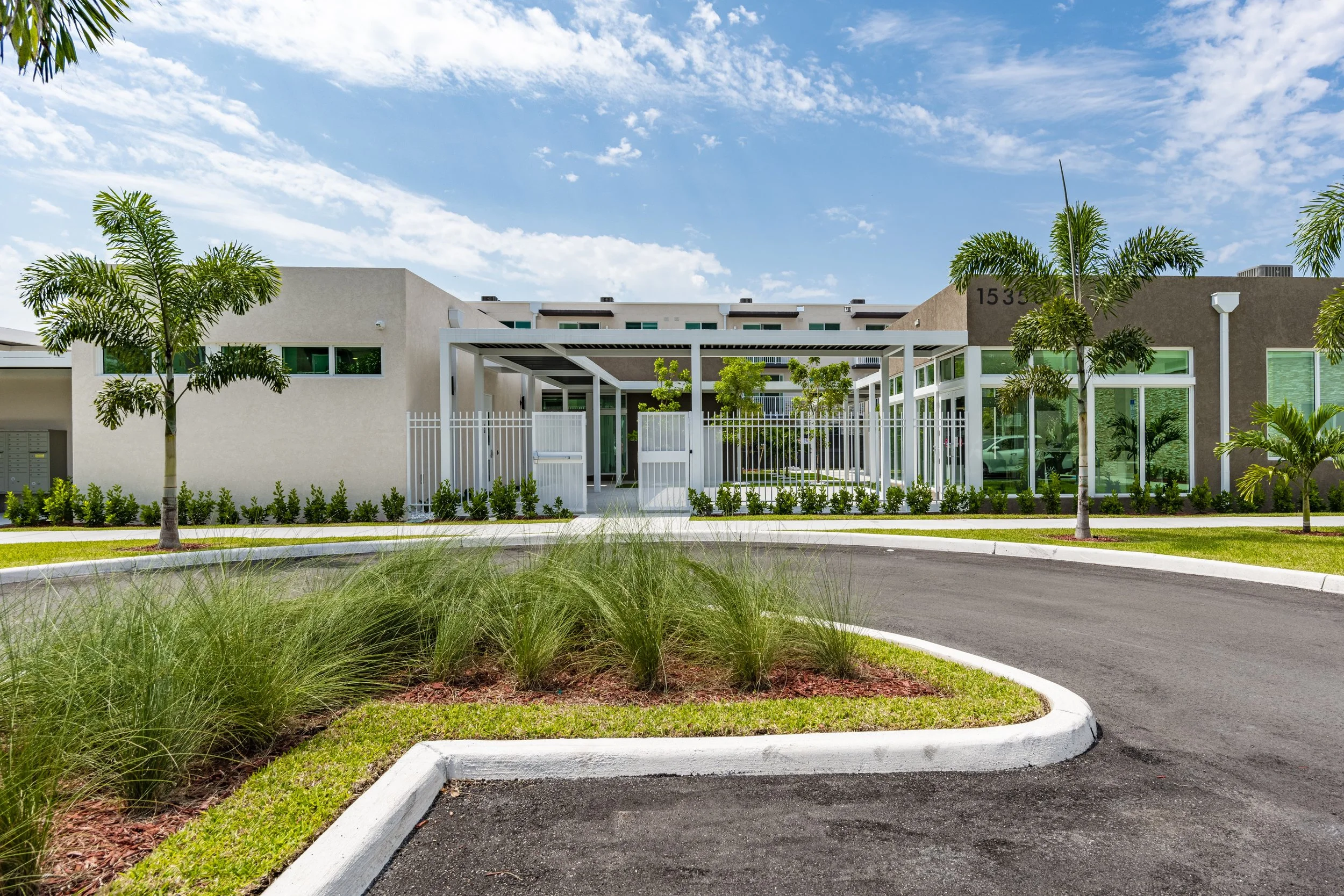 Modern apartment building with landscaped lawn and palm trees in front, paved driveway curving around a grassy island, blue sky with clouds.