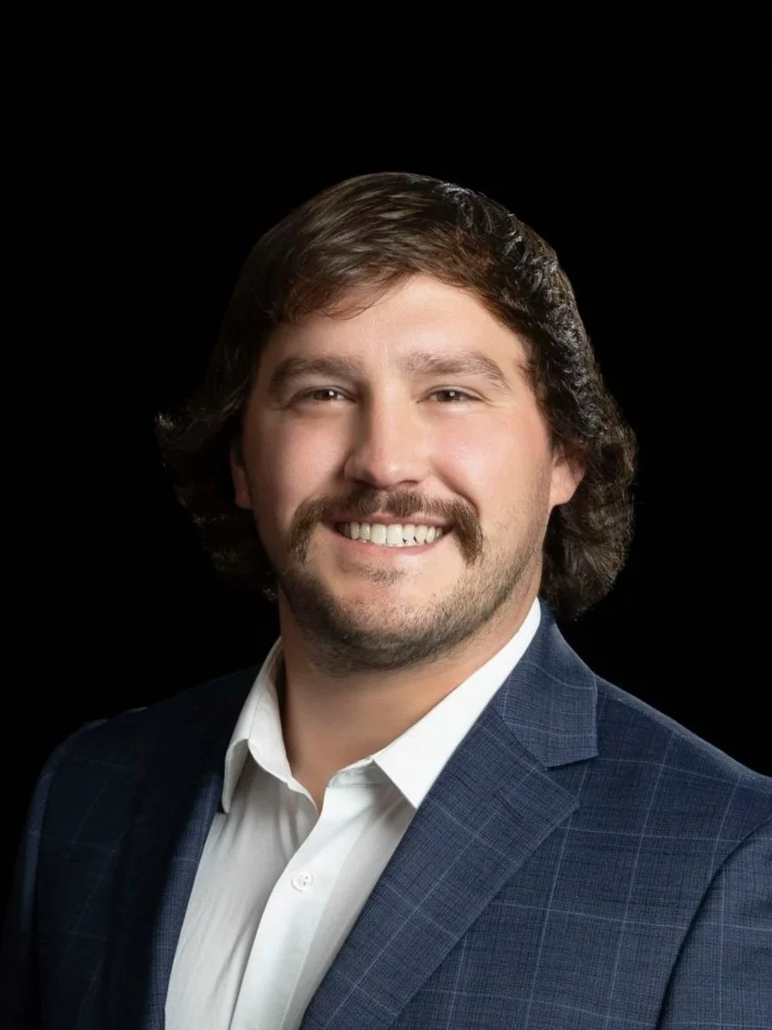 A smiling man with short brown hair wearing a plaid shirt against a dark gray background.