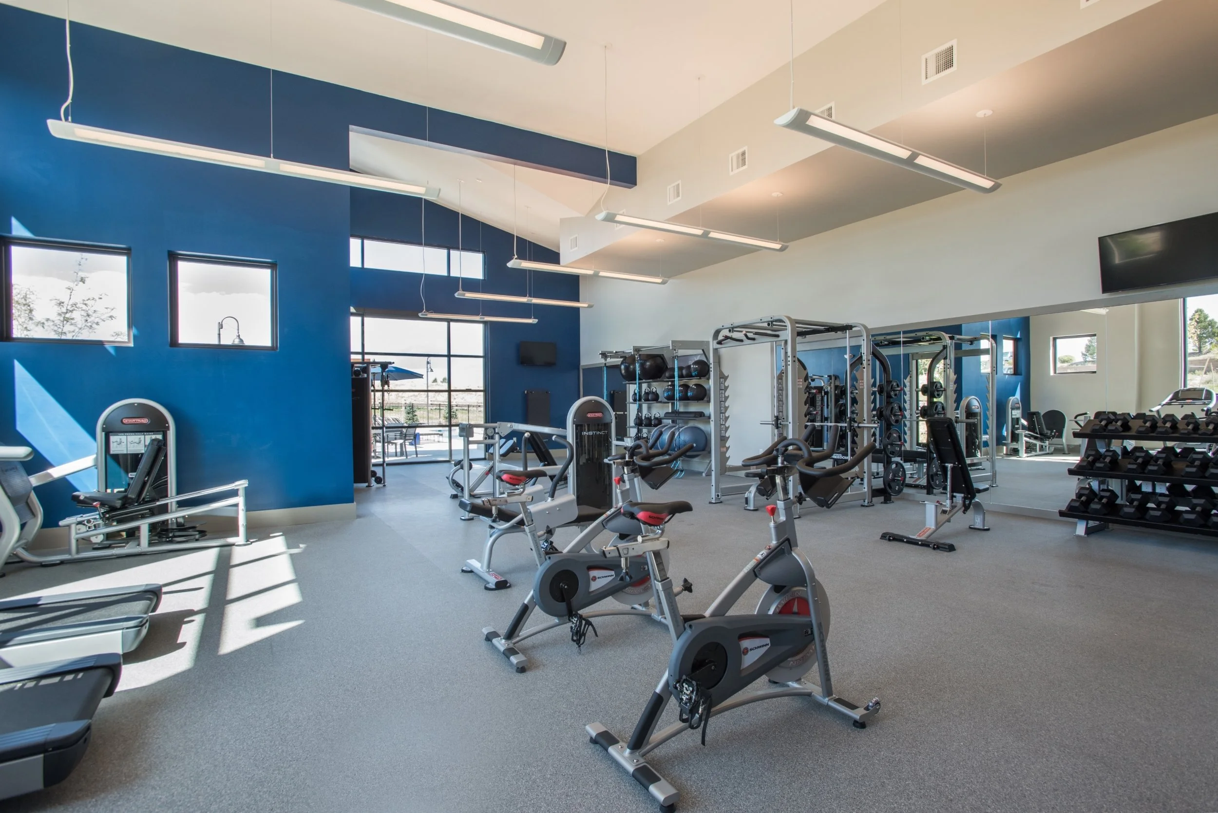 Modern gym with exercise bikes, weight rack, and fitness equipment inside a bright room with large windows, blue and white walls, and ceiling lights.