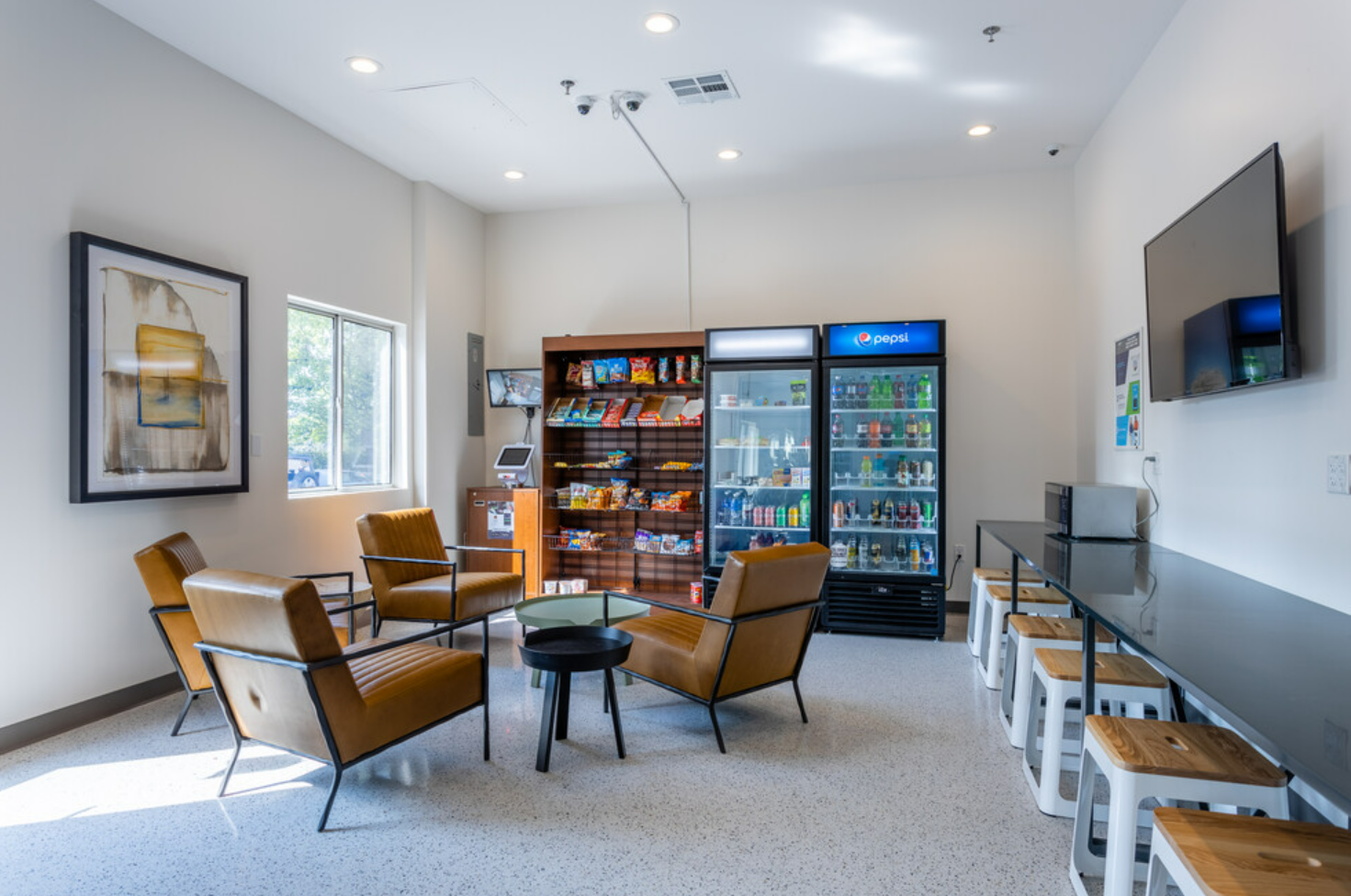A modern waiting area with four brown armchairs arranged around a small black table, a glass coffee table, a snack and beverage vending machine, a refrigerator, a wall-mounted TV, and a long countertop with white stools. A window on the left wall let