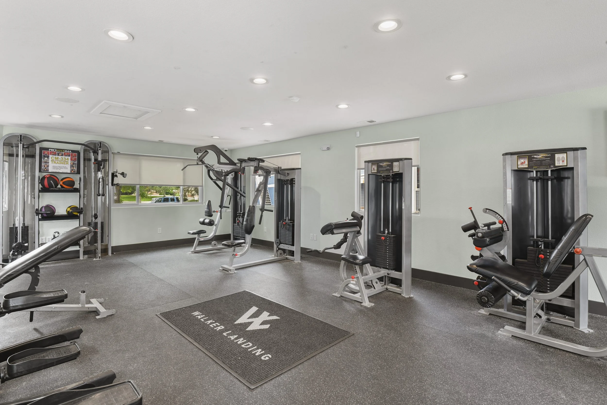 A modern gym with various fitness equipment including weight machines, a seated leg press, and a multi-station workout machine, with windows letting in natural light.