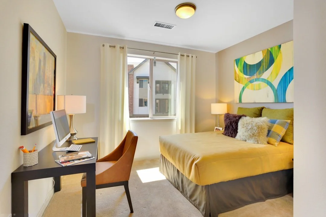 A cozy bedroom with a bed, colorful pillows, a window with curtains, a desk with a computer, and modern artwork on the walls.