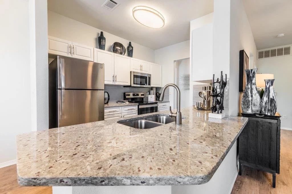 Modern kitchen with white cabinets, stainless steel refrigerator, microwave, and oven. Granite countertop island with double sink and faucet, decorative vases and sculptures.