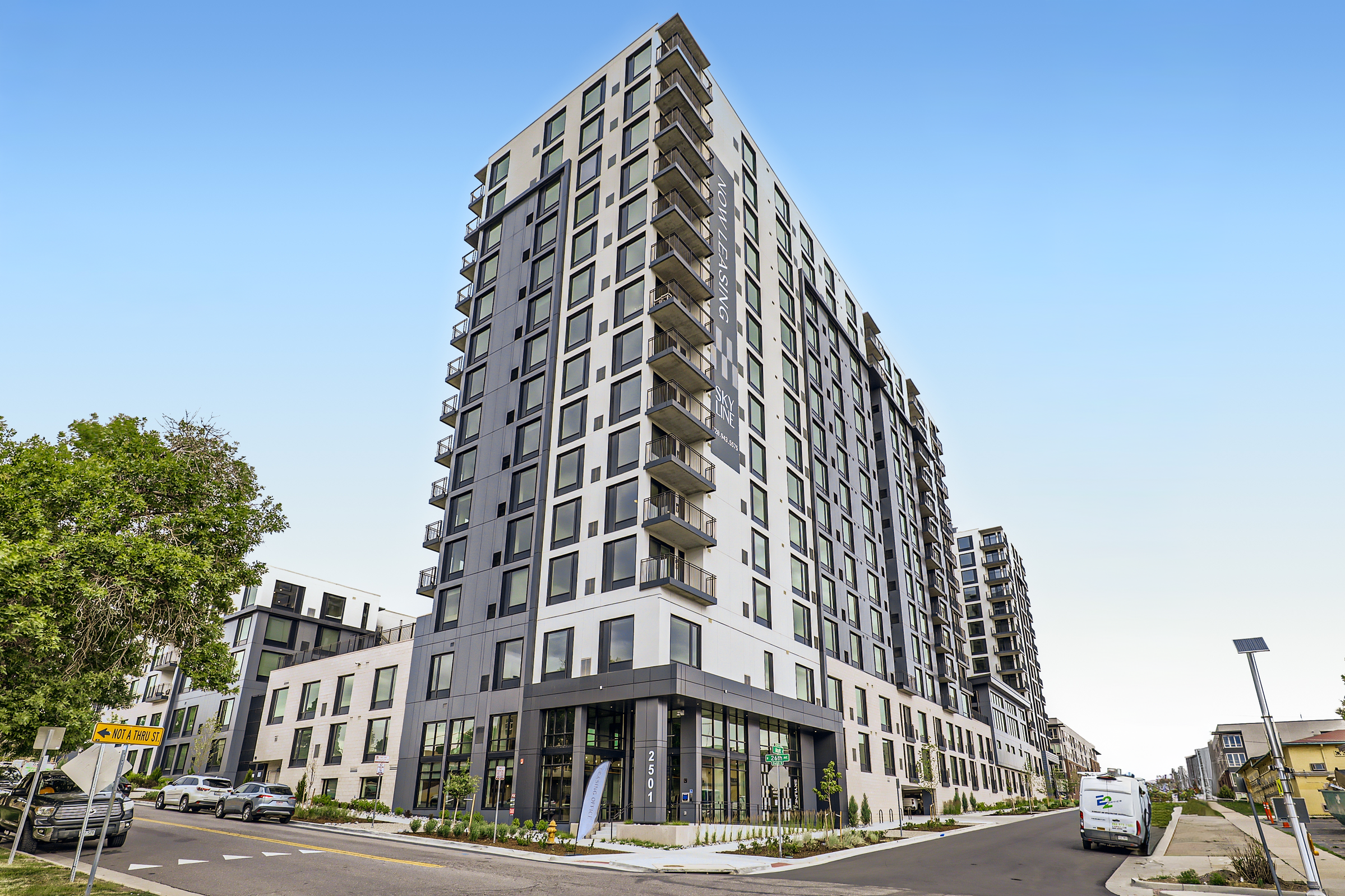 Modern multi-story residential building with glass windows and balconies, located at an urban street corner on a sunny day.