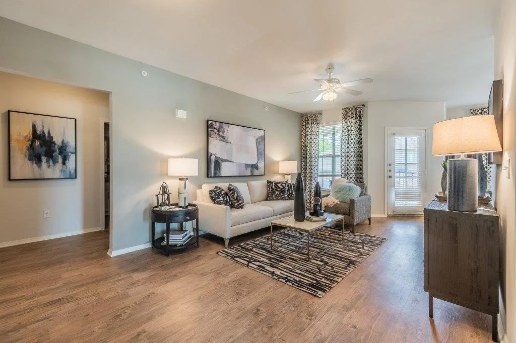 Living room with a white sofa, armchair, coffee table, wall art, table lamps, curtains, and hardwood flooring.
