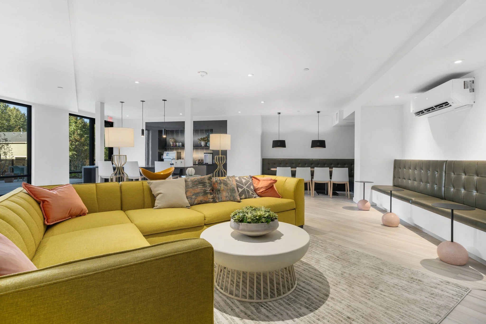 Modern living room with yellow sectional sofa, multicolored pillows, white coffee table with a plant, light wood flooring, and large windows. In the background, there are dining areas with white chairs and hanging pendant lights.