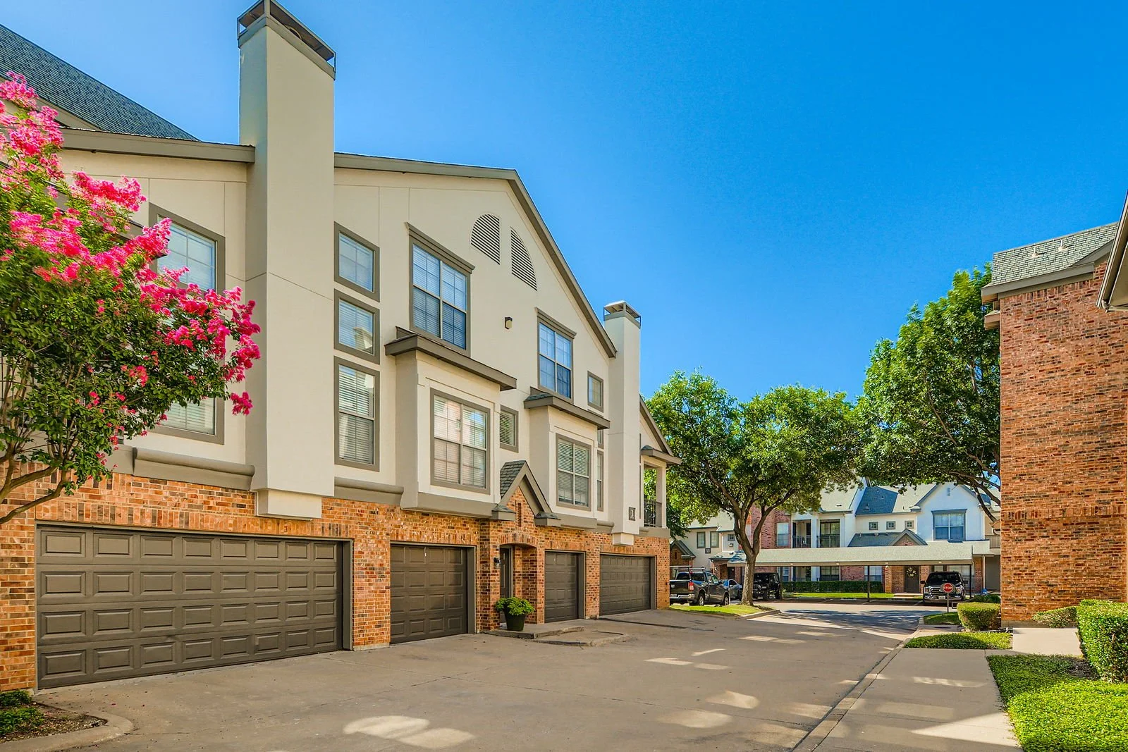A residential courtyard with multiple three-story townhomes with garages, trees, and parked cars under a clear blue sky.