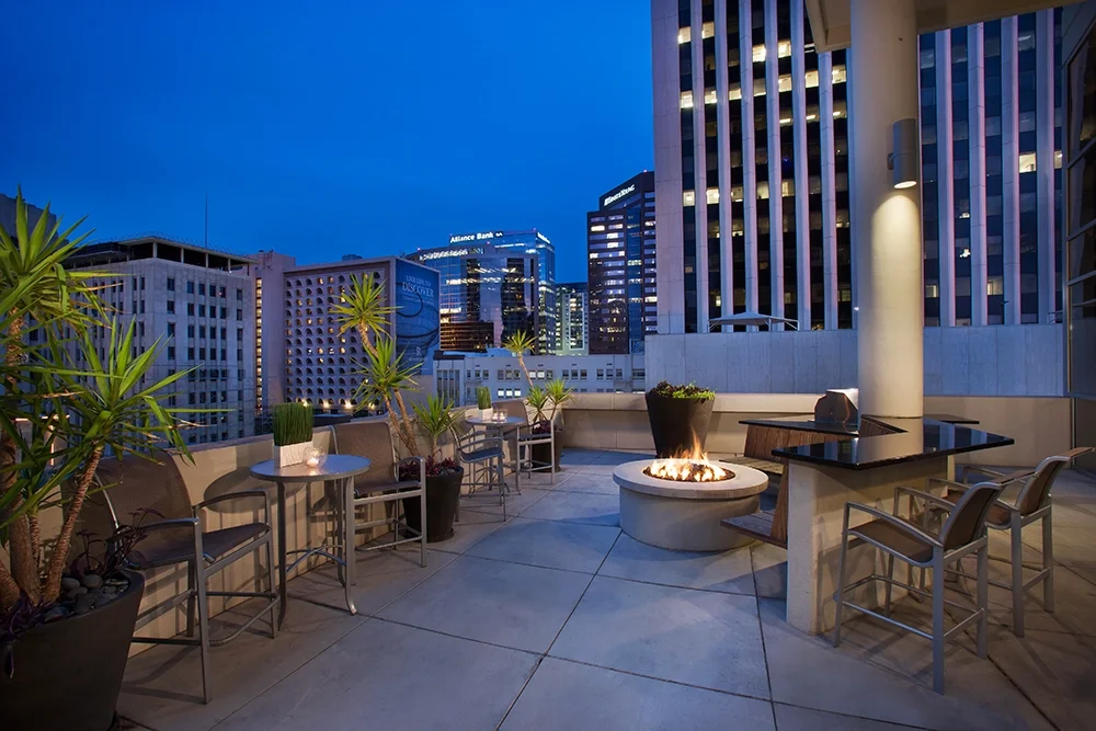 Outdoor rooftop patio at dusk with modern furniture, potted plants, a fire pit, and city skyline in the background.