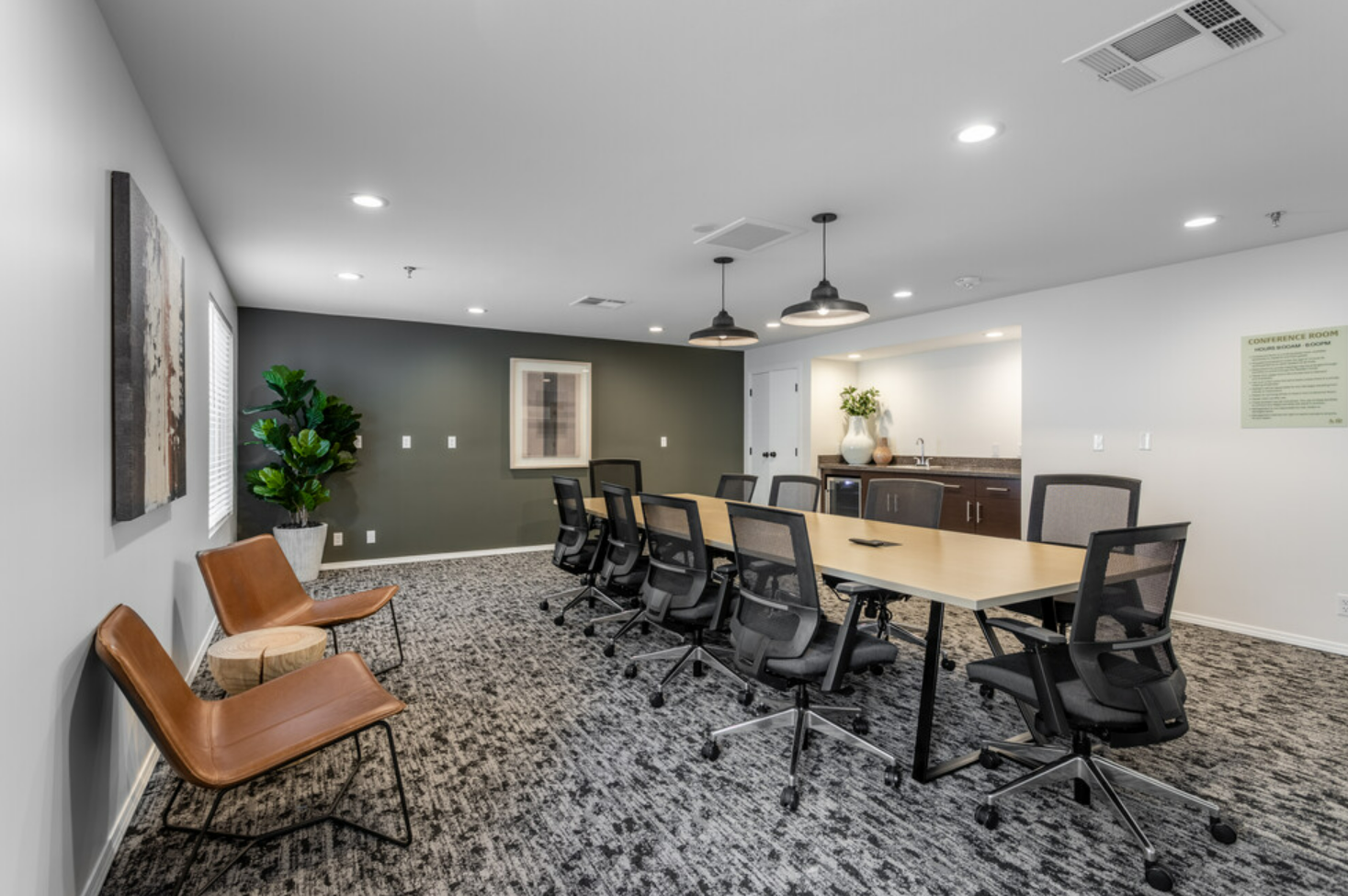 Conference room with a long table, black chairs, and a small seating area with two leather chairs and a side table, decorated with artwork, indoor plants, and pendant lighting.