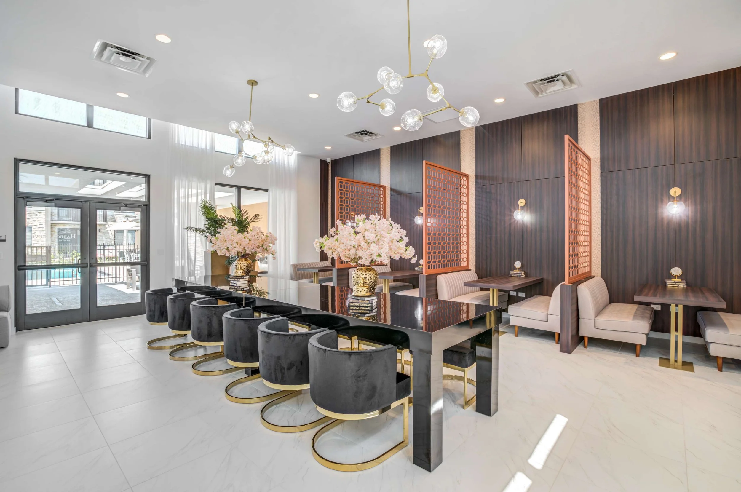 Modern hotel lobby with black marble table, gray velvet chairs with gold bases, pink floral centerpieces, wooden wall panels, and hanging modern light fixtures.