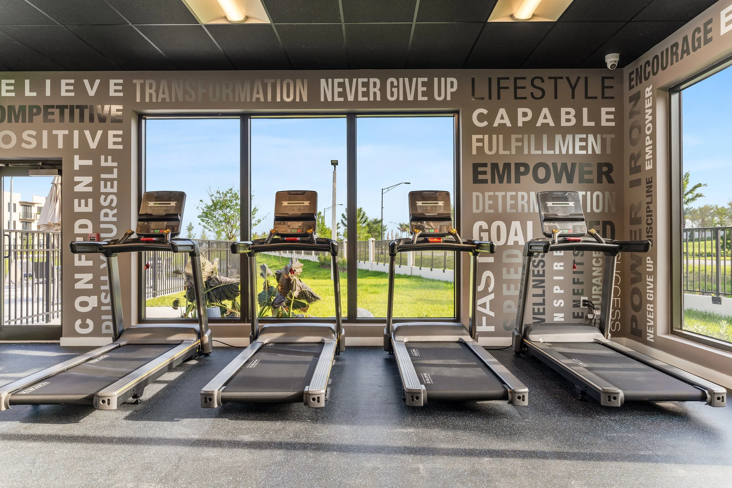 Four treadmills positioned side by side in a gym with a large window showing outdoors.