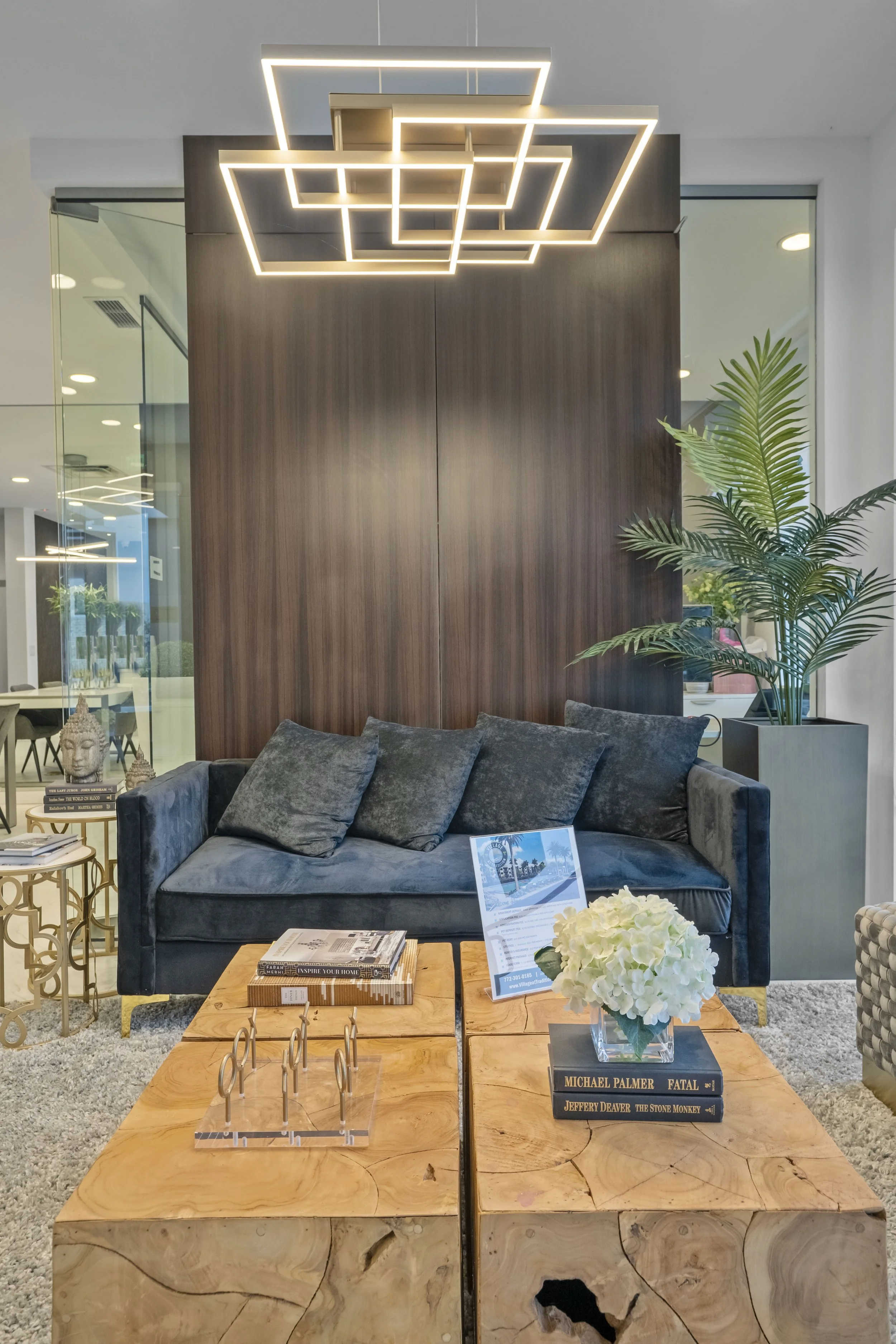 Modern lobby with wooden coffee tables, black velvet sofa, green plant, and geometric ceiling lights.