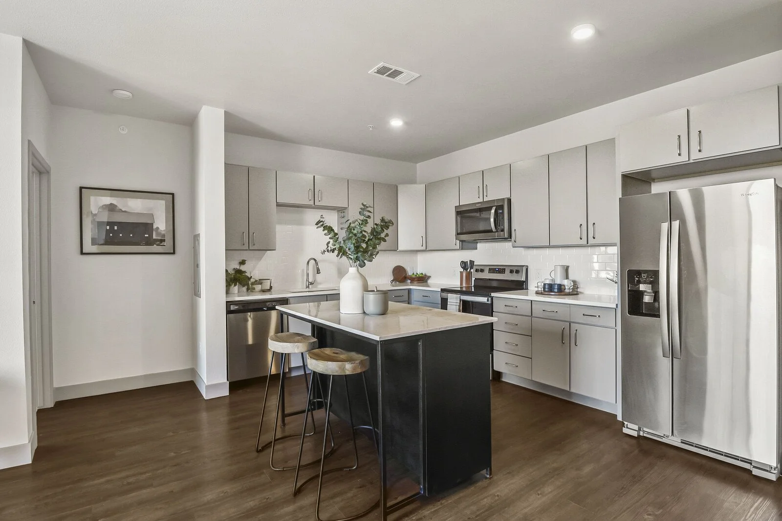Modern kitchen with white cabinets, stainless steel refrigerator, oven, microwave, and a central island with two bar stools, decorated with a vase of greenery.