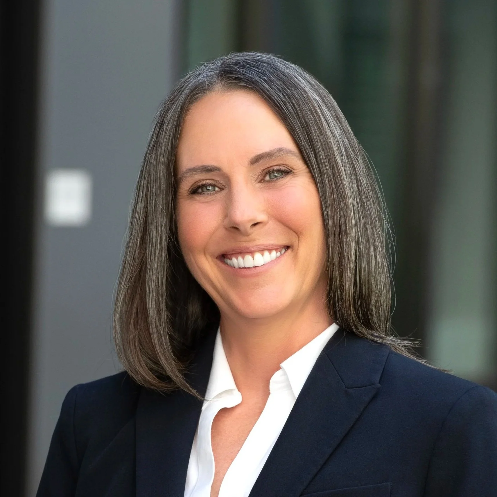 Close-up of a smiling woman with shoulder-length brown hair, wearing a navy blazer and white shirt, in an outdoor or office setting.