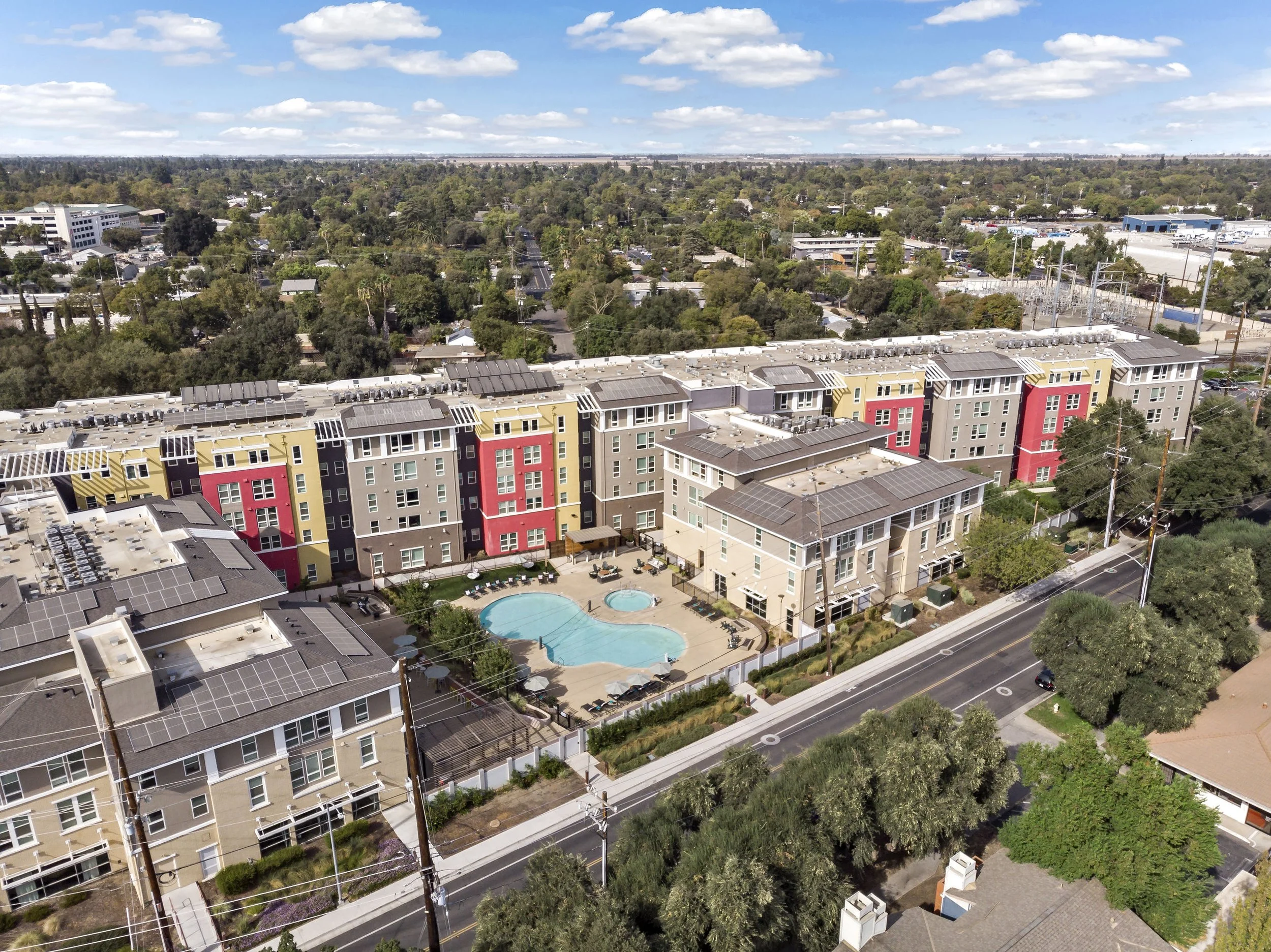 Aerial view of a modern apartment complex with a swimming pool, surrounded by trees and a street, under a partly cloudy sky.