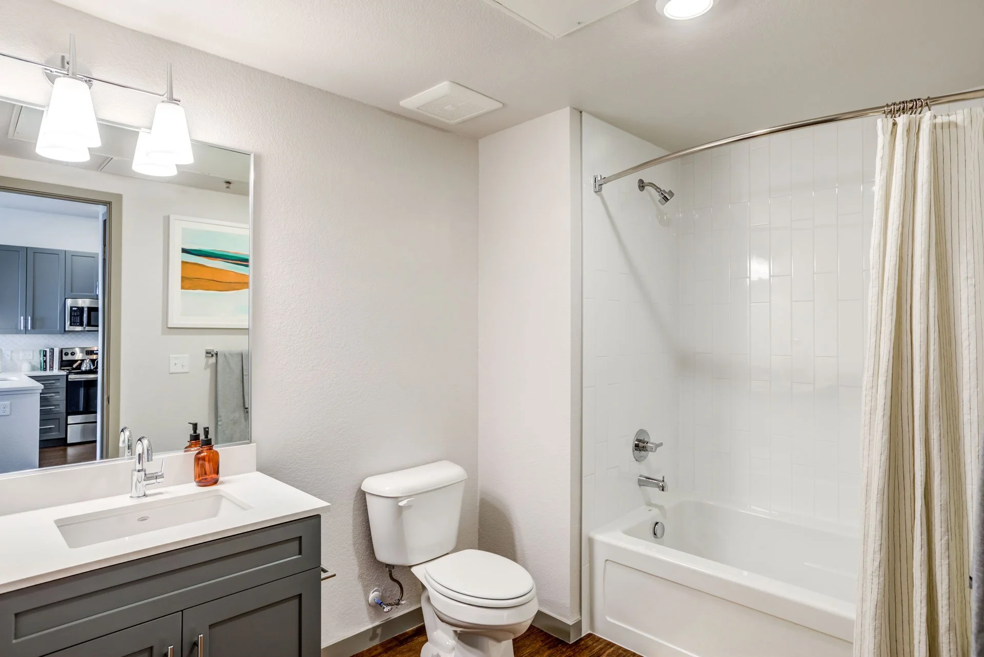 Bathroom with a white toilet, a vanity with a sink and a mirror, and a bathtub/shower combo with a curtain. There is a soap dispenser on the vanity.