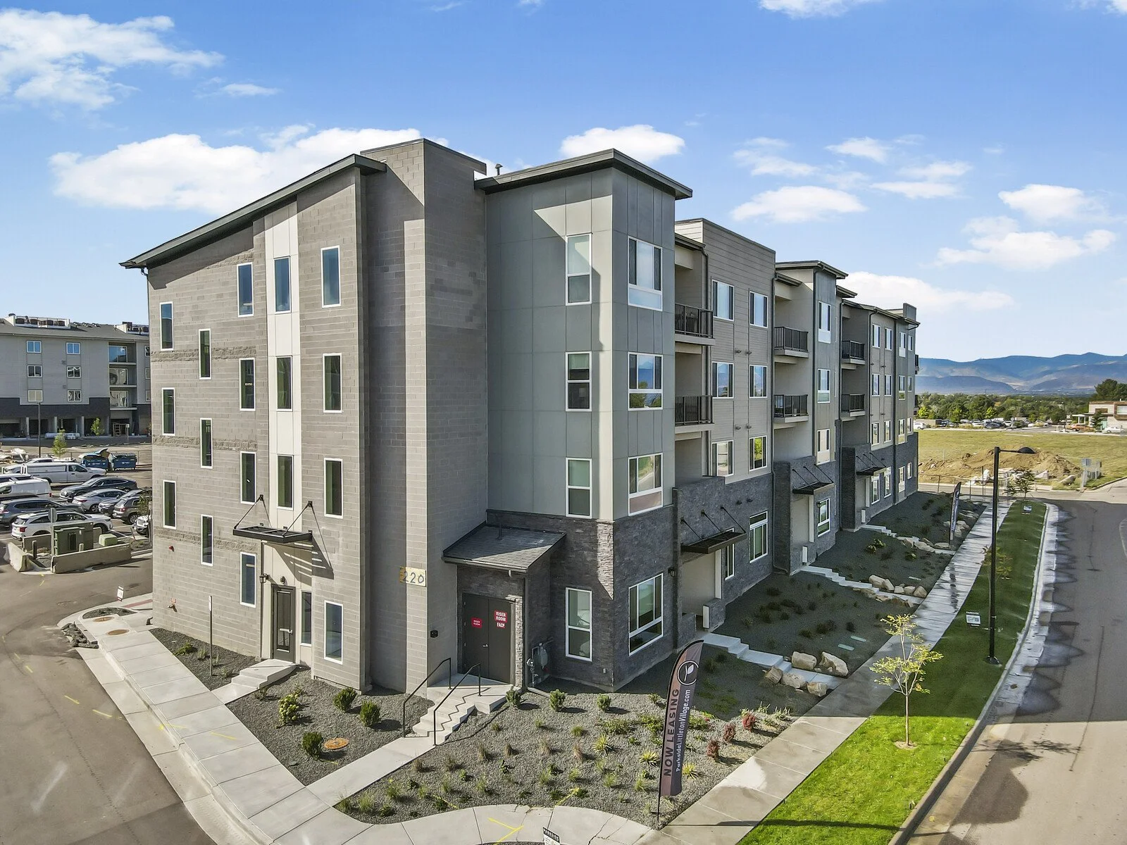 Modern multi-story apartment building with gray and beige exterior, multiple balconies, windows, and a parking lot in the foreground.