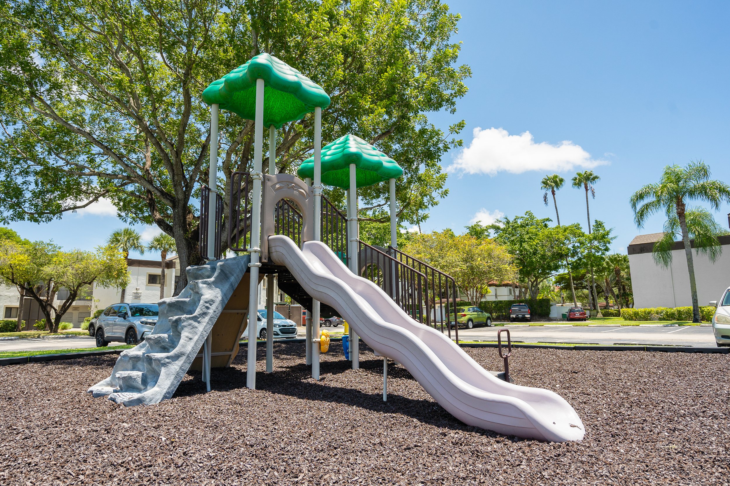 Children's playground with slides and climbing wall, surrounded by trees and parked cars on a sunny day.