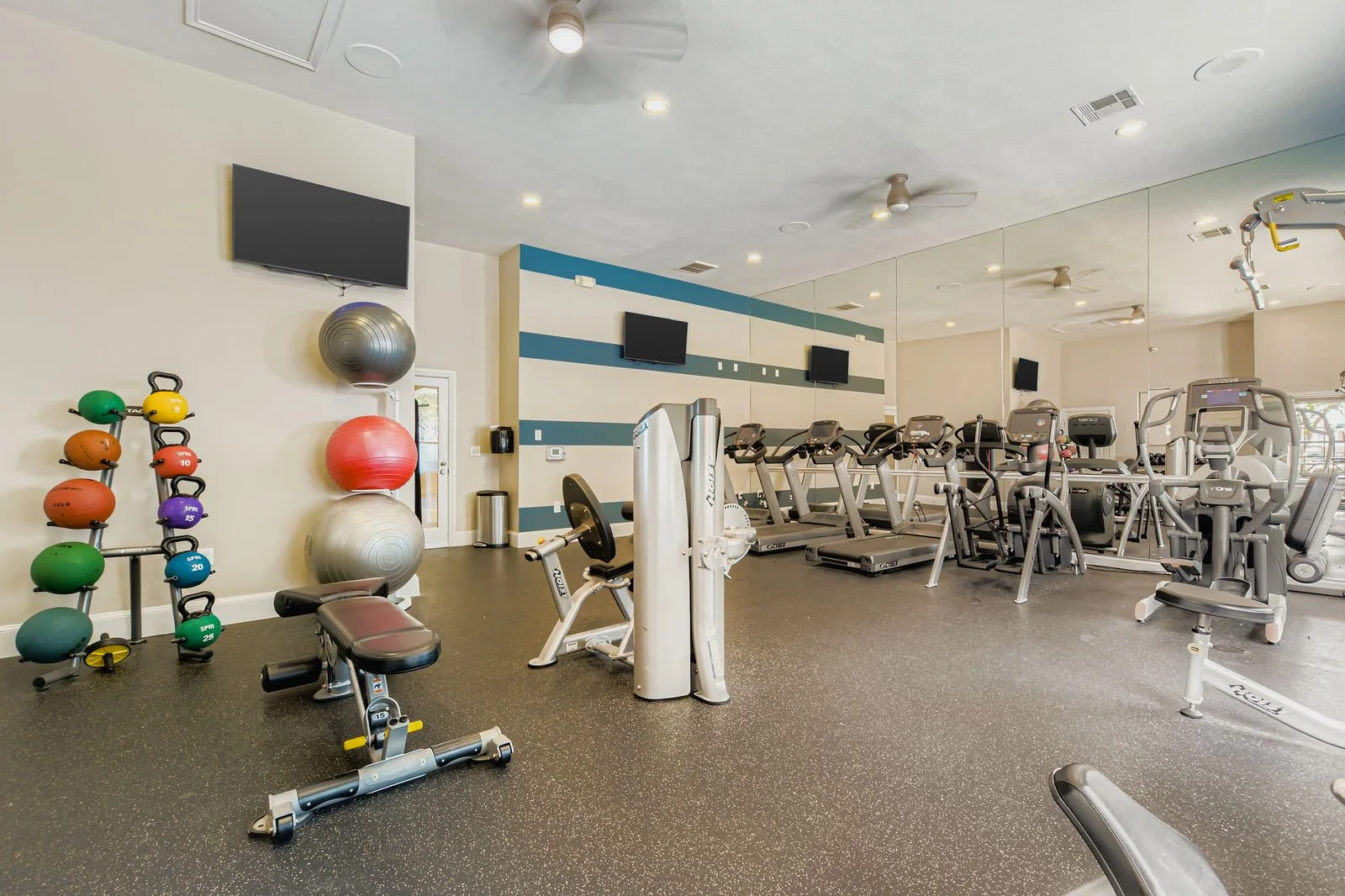 Empty gym with various exercise equipment, including treadmills, elliptical machines, weight benches, and colorful kettlebells.