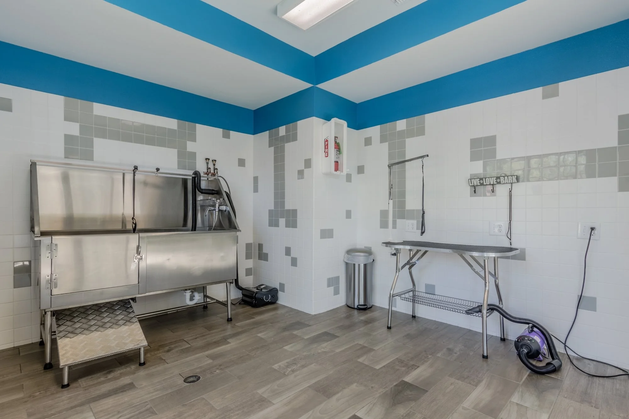 An empty pet grooming station with stainless steel sinks, grooming table, and cleaning supplies in a tiled room.