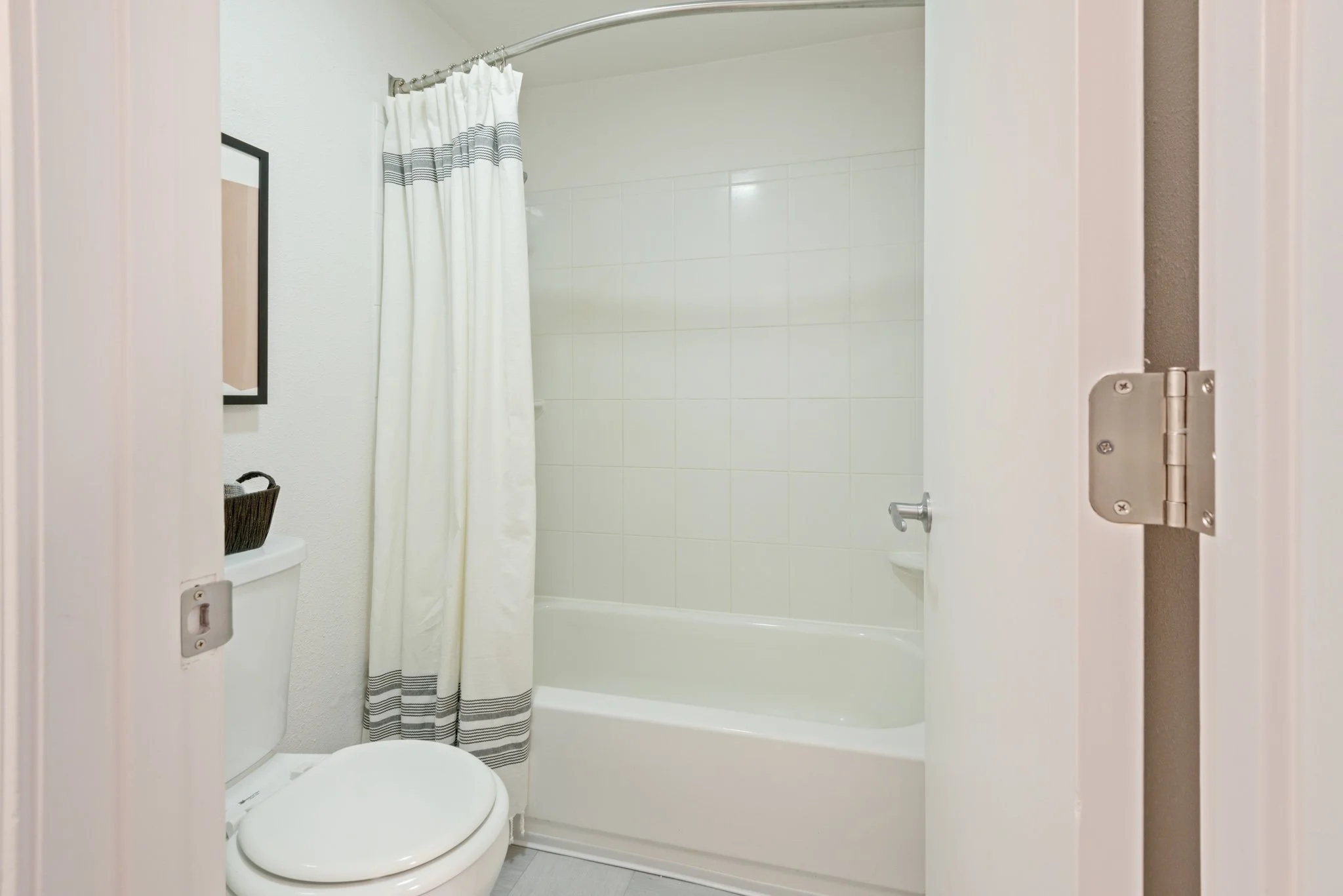 Inside a small bathroom with a white toilet and bathtub, a cream shower curtain with gray stripes, white tiled walls, a partial view of a framed picture and a small black basket on the toilet tank.