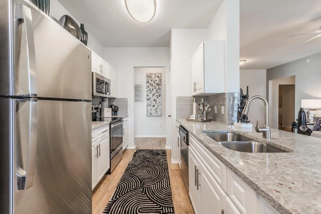 Modern kitchen with stainless steel refrigerator, microwave, and oven, white cabinets, granite countertop, double sink, beige backsplash, black and white patterned rug, and various kitchen utensils.