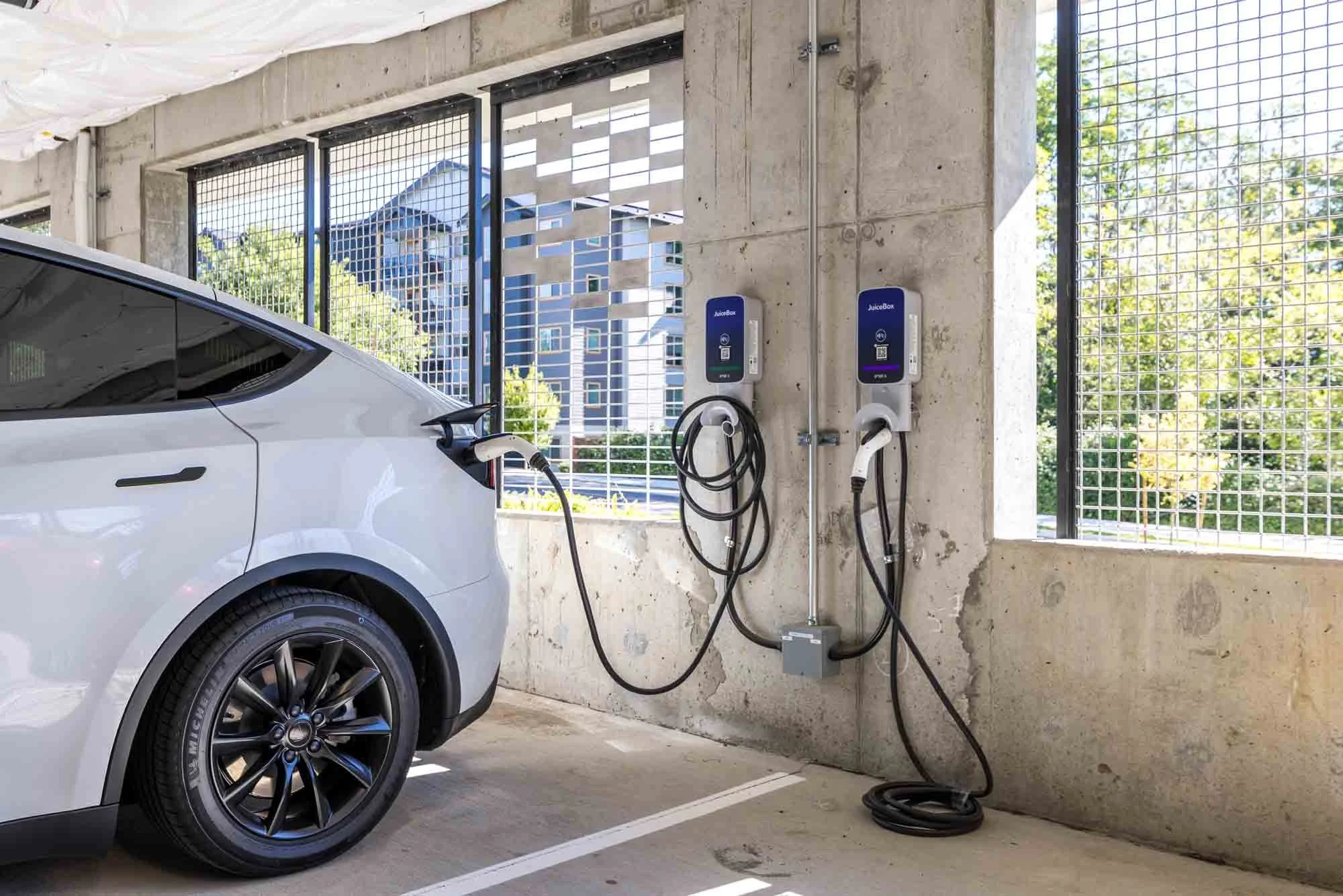 White electric vehicle charging at a station in a parking garage with metal grid windows and a cityscape view.