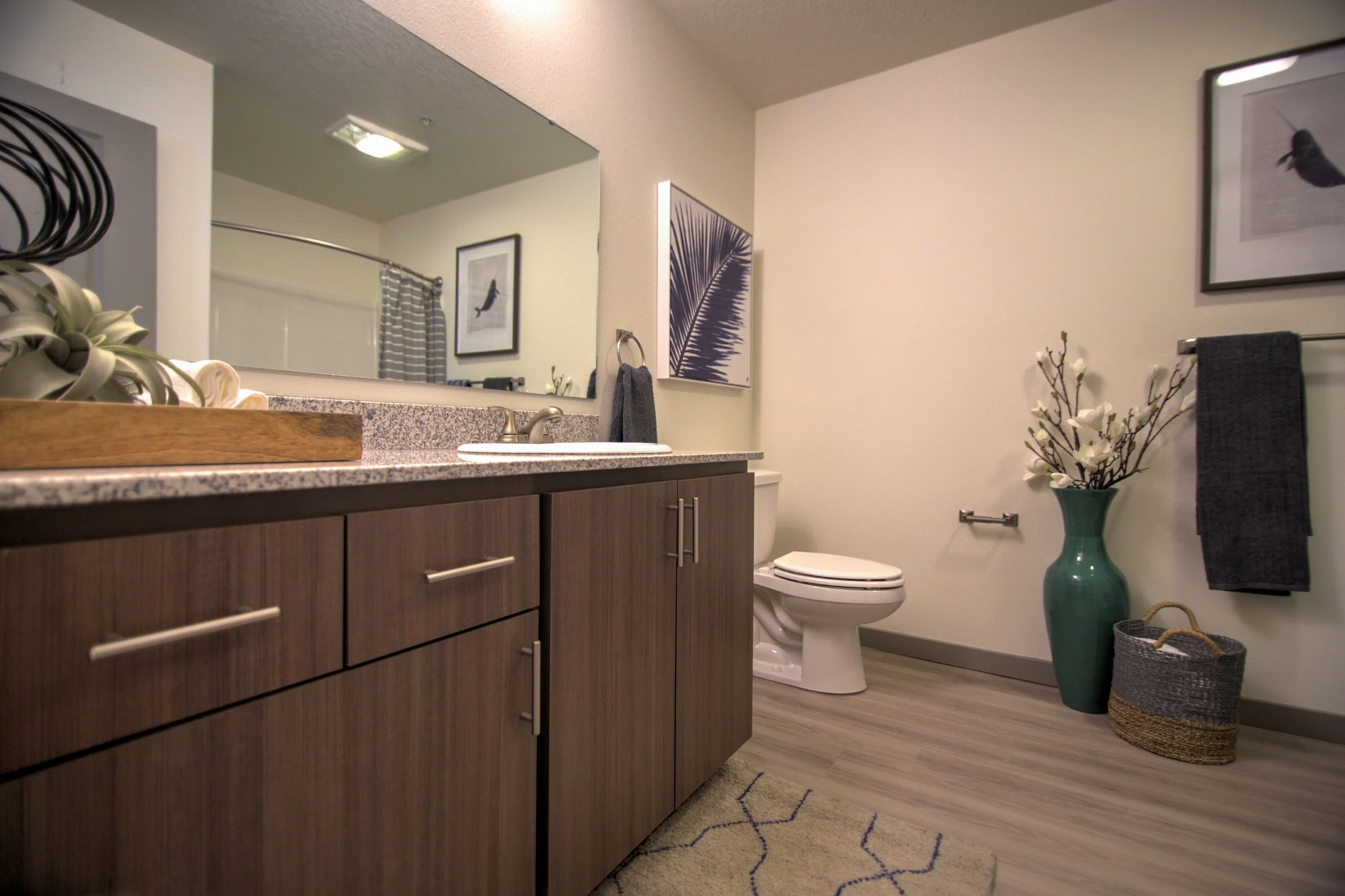 A modern bathroom featuring a wooden vanity with a granite countertop, a large wall mirror, a white toilet, decorative art on the walls, a green vase with white flowers, a gray towel, and a woven basket on a light wood floor.