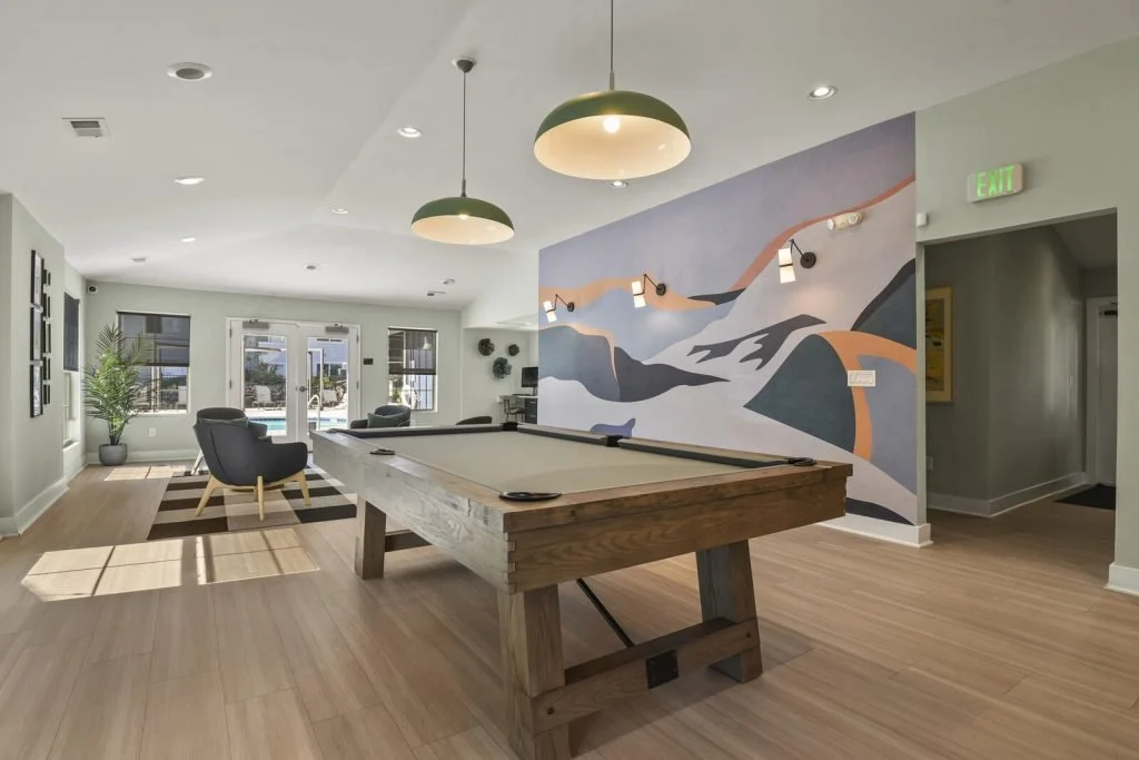 Interior of a recreational room with a pool table, lounge chairs, a potted plant, large windows, and a colorful abstract mural on the wall.