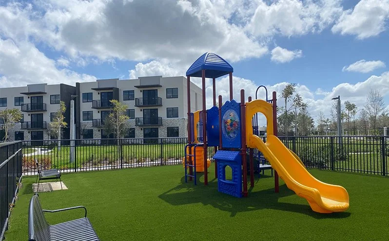 Colorful playground with a yellow slide and blue and red structures, surrounded by a black fence, with apartment buildings and cloudy sky in the background.