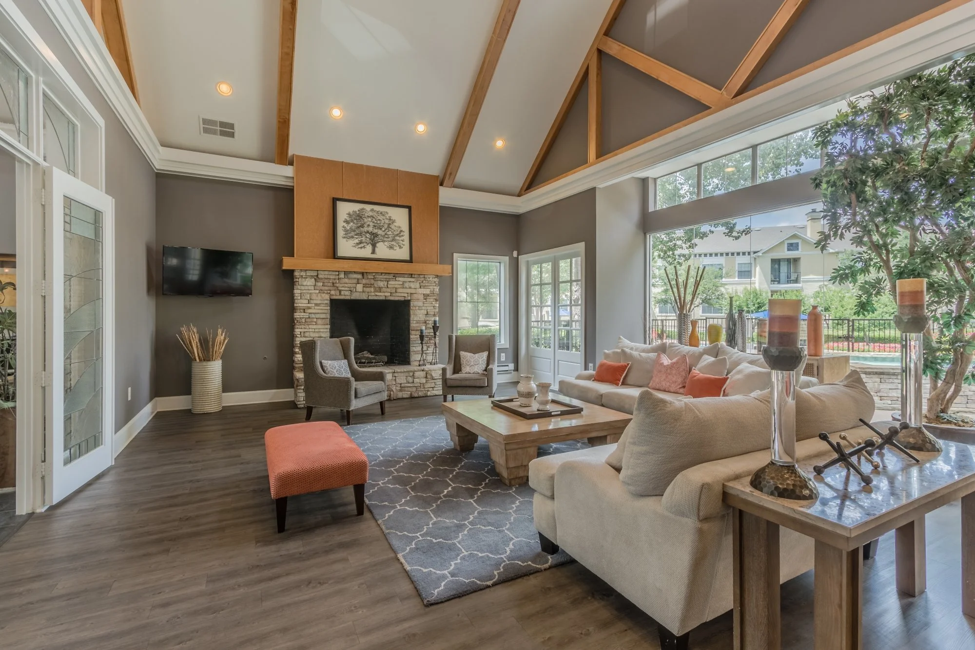 Living room with fireplace, large windows, gray walls, wooden ceiling beams, beige sofa with orange and pink pillows, two gray armchairs, a coffee table, a patterned rug, and decorative candles and vases.