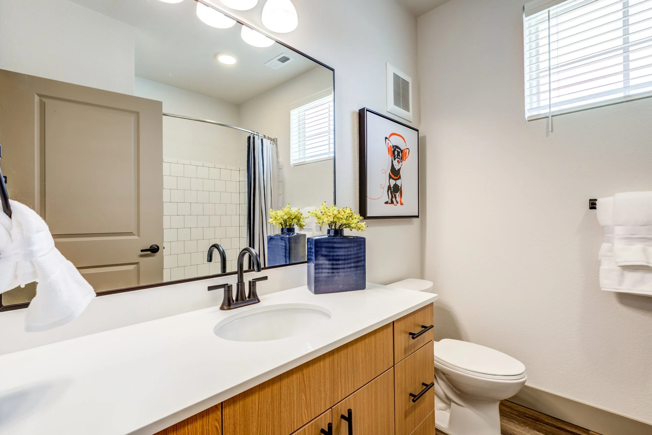 Modern bathroom with wooden vanity, white countertop, oval sink, large mirror, black faucet, potted flowers, framed art of a dog with headphones, window with blinds, shower with white tiles, white towels, and beige walls.