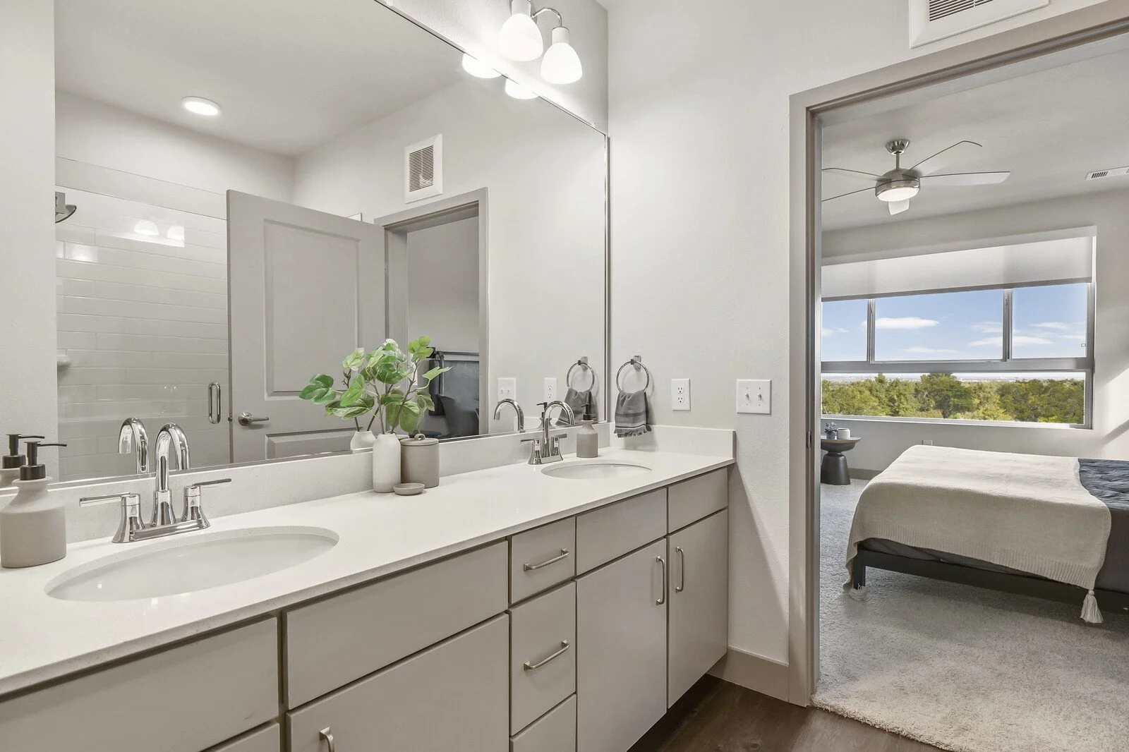A bathroom vanity with two sinks, a large mirror, decorative vases with green plant, soap dispensers, and gray towels on rings. Behind the vanity, a door leads to a shower with white subway tile. To the right, an open doorway reveals a bedroom with a