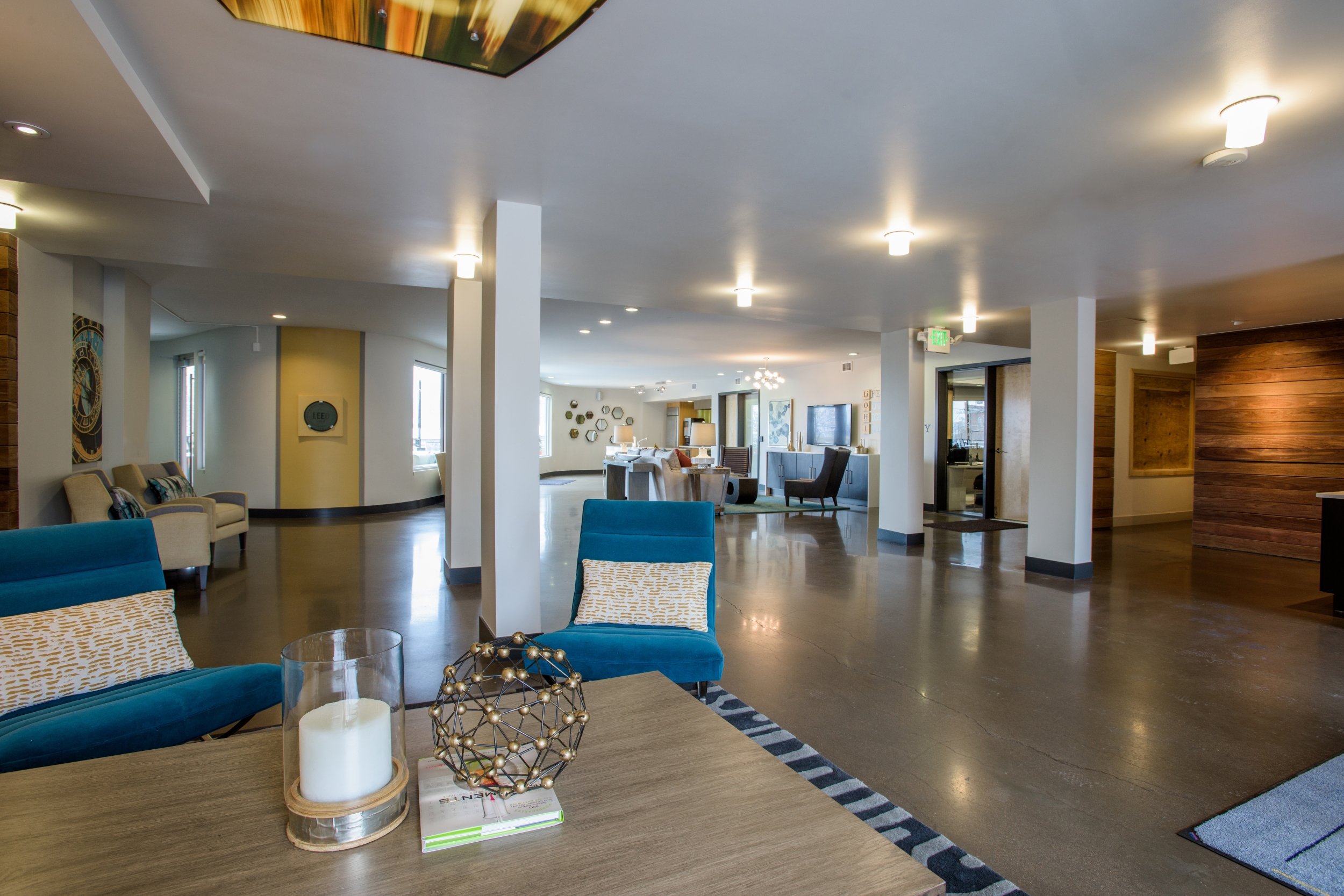 Modern hotel lobby with a wooden table in the foreground holding a candle, a decorative globe, and a magazine. The lobby features blue and beige chairs, a white pillar, and a polished wooden floor. There are multiple lights on the ceiling and various
