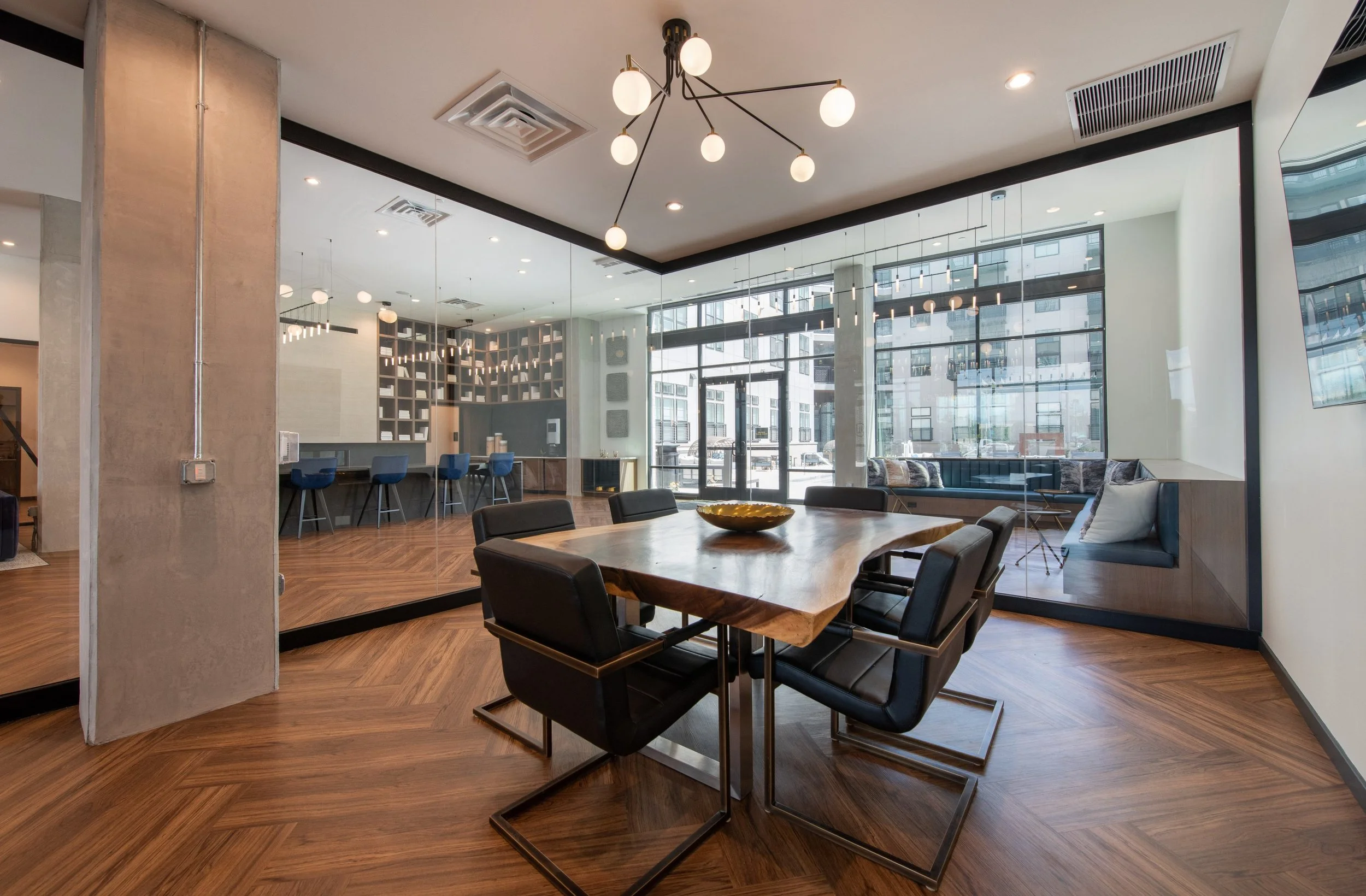 Modern interior office space with a wooden dining table, black chairs, a view of a bar area with three blue bar stools, and large windows letting in natural light.