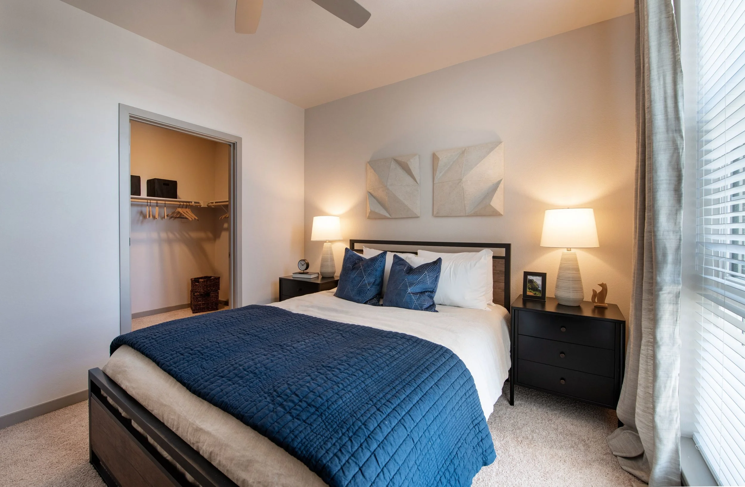 Bedroom with a bed, dark headboard, white bedding, blue quilt and pillows, two black nightstands with lamps and decor, open closet with hanging clothes, window with blinds and curtains, ceiling fan.