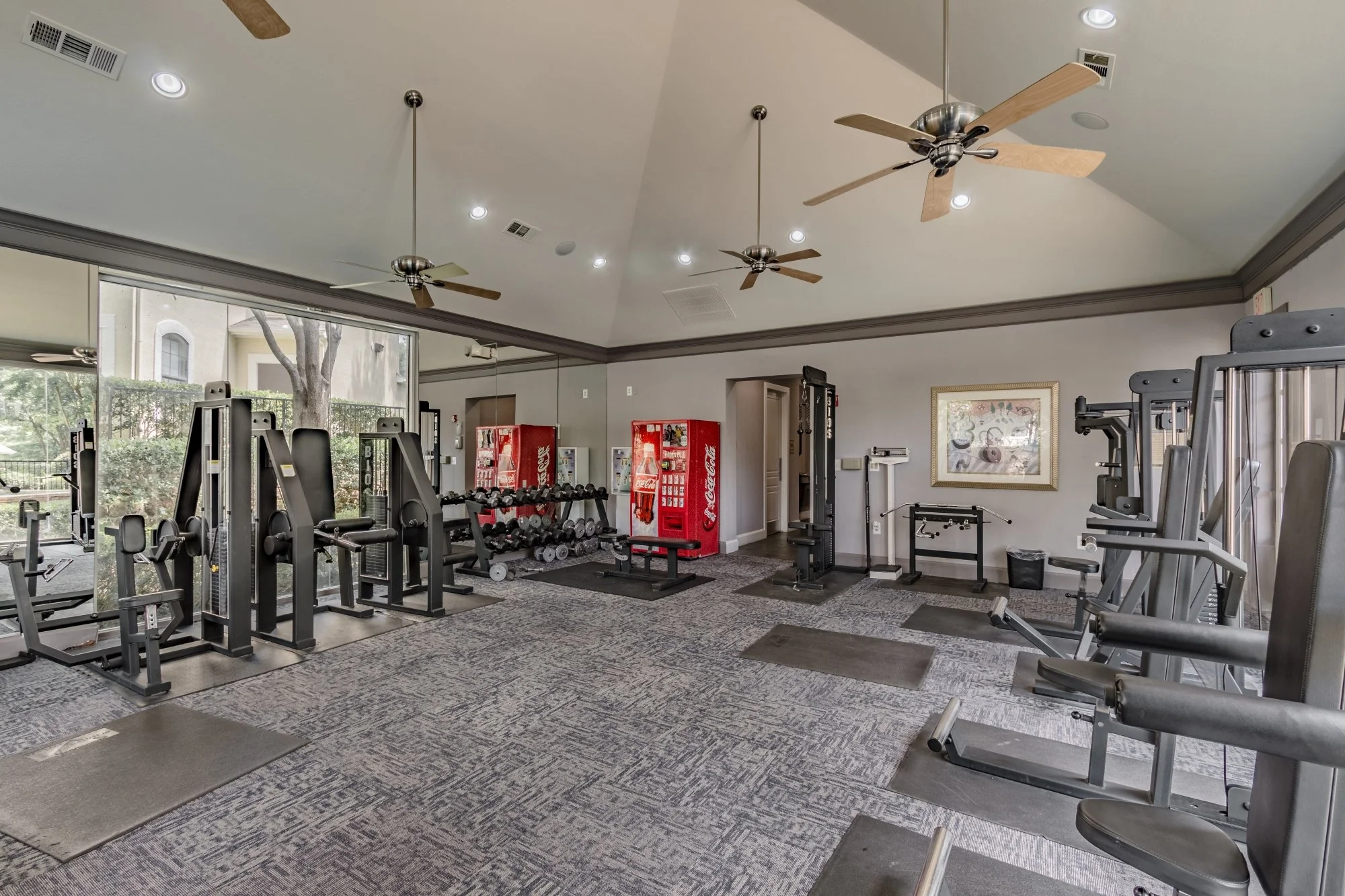 Empty gym with various workout equipment including weight machines, free weights, and vending machines. Large window along one wall with ceiling fans and lights above.