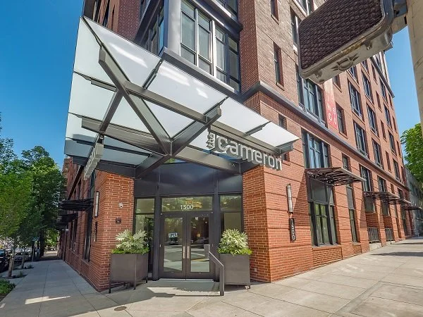Exterior view of a modern brick apartment building named The Cameron, with glass doors and a canopy at the entrance, along a city sidewalk with planters and trees.