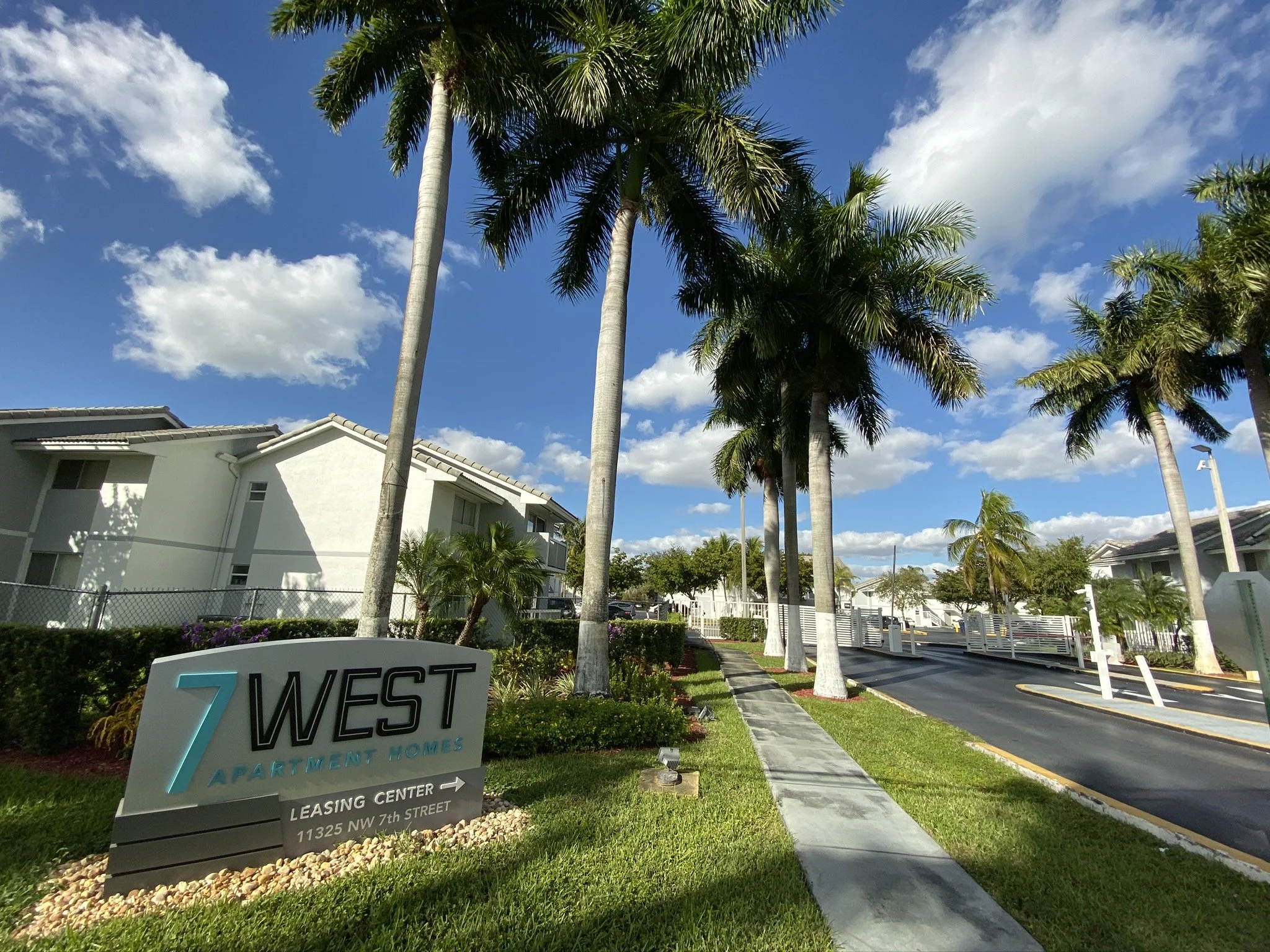 A sign for 7 West Apartment Homes in front of a residential complex with palm trees, a sidewalk, and a street under a blue sky with clouds.