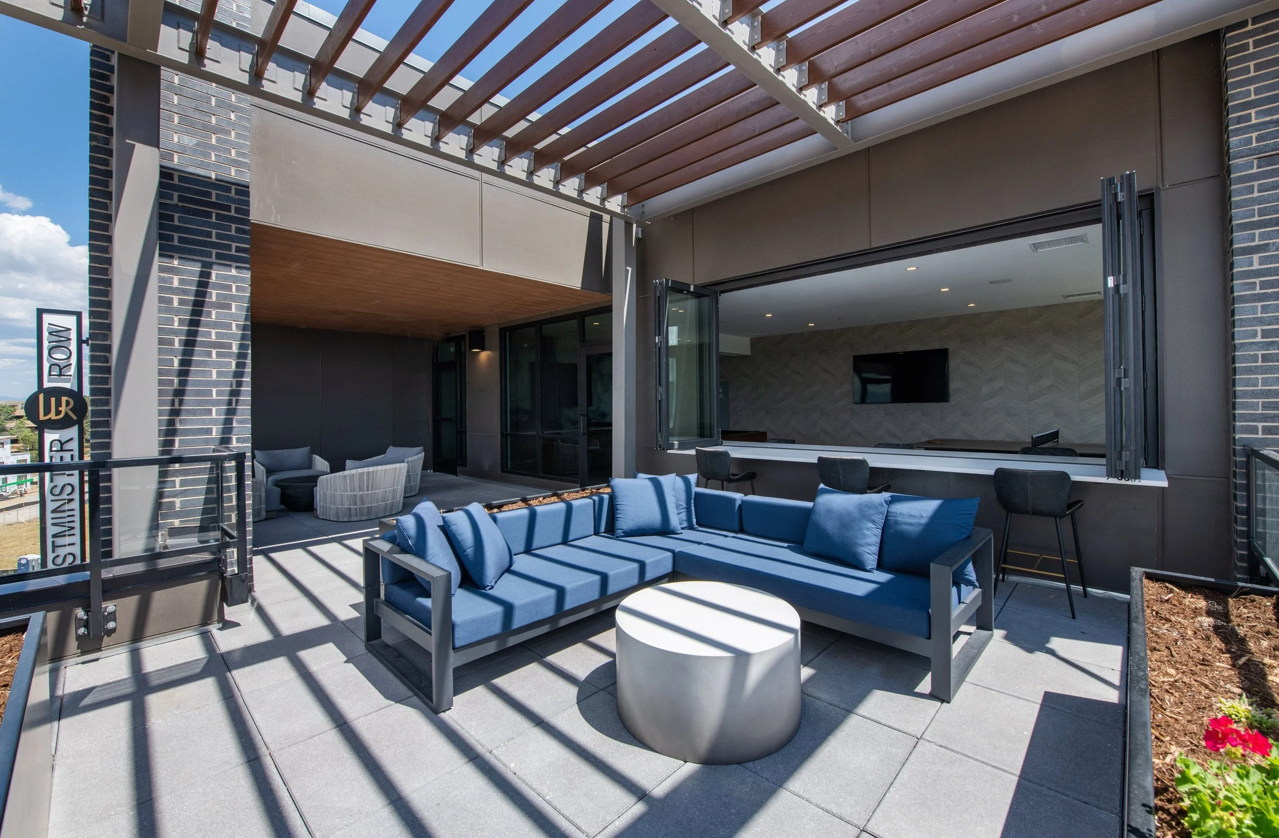 Outdoor rooftop patio with blue cushioned sectional sofa, gray side table, black outdoor bar stools, and a view of the city sky.