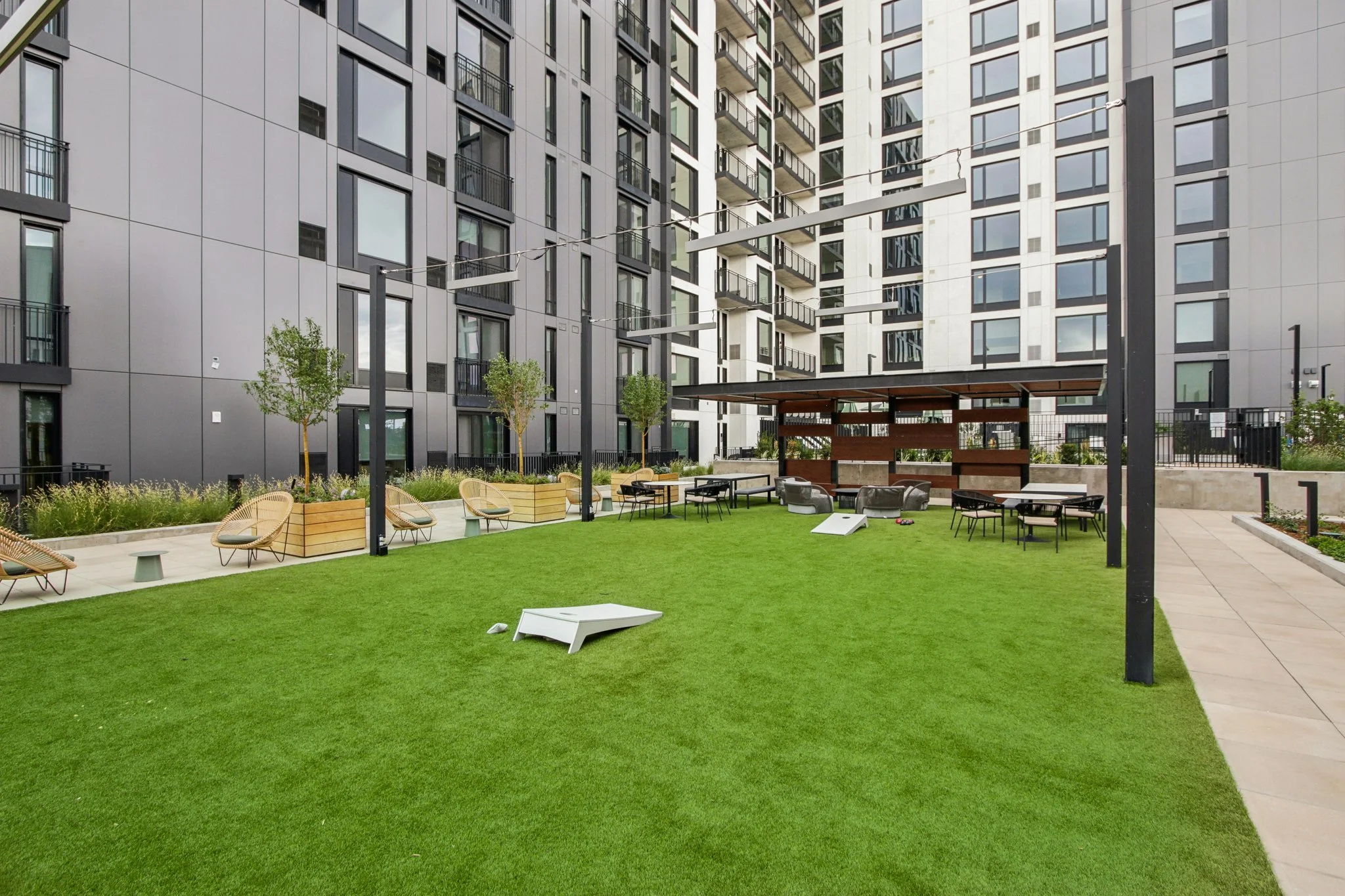 An outdoor communal area with green grass, lounge chairs, tables, and a bar area, surrounded by tall modern apartment buildings.