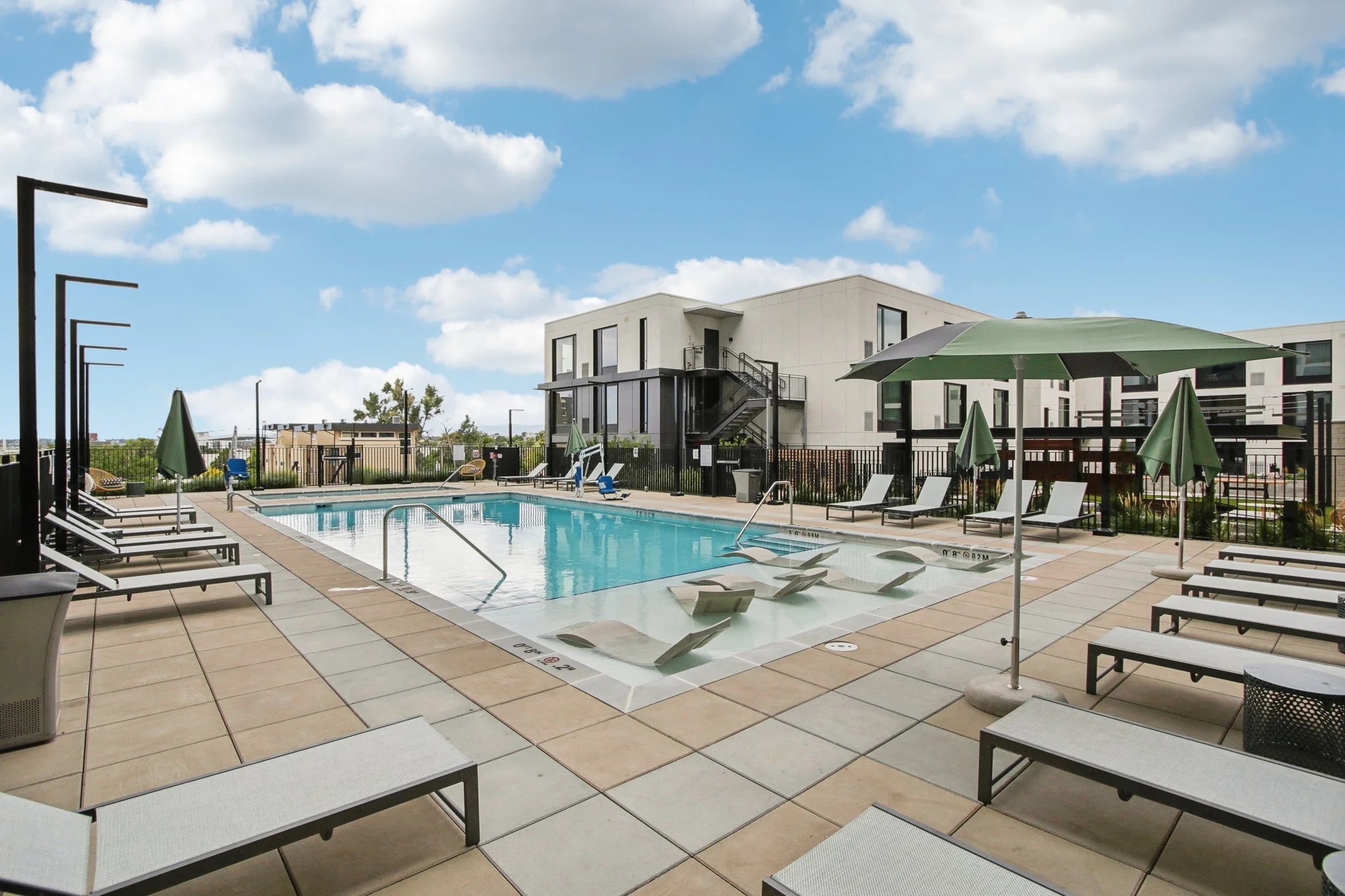 An outdoor swimming pool area with lounge chairs, umbrellas, and a modern residential building in the background, under a partly cloudy sky.
