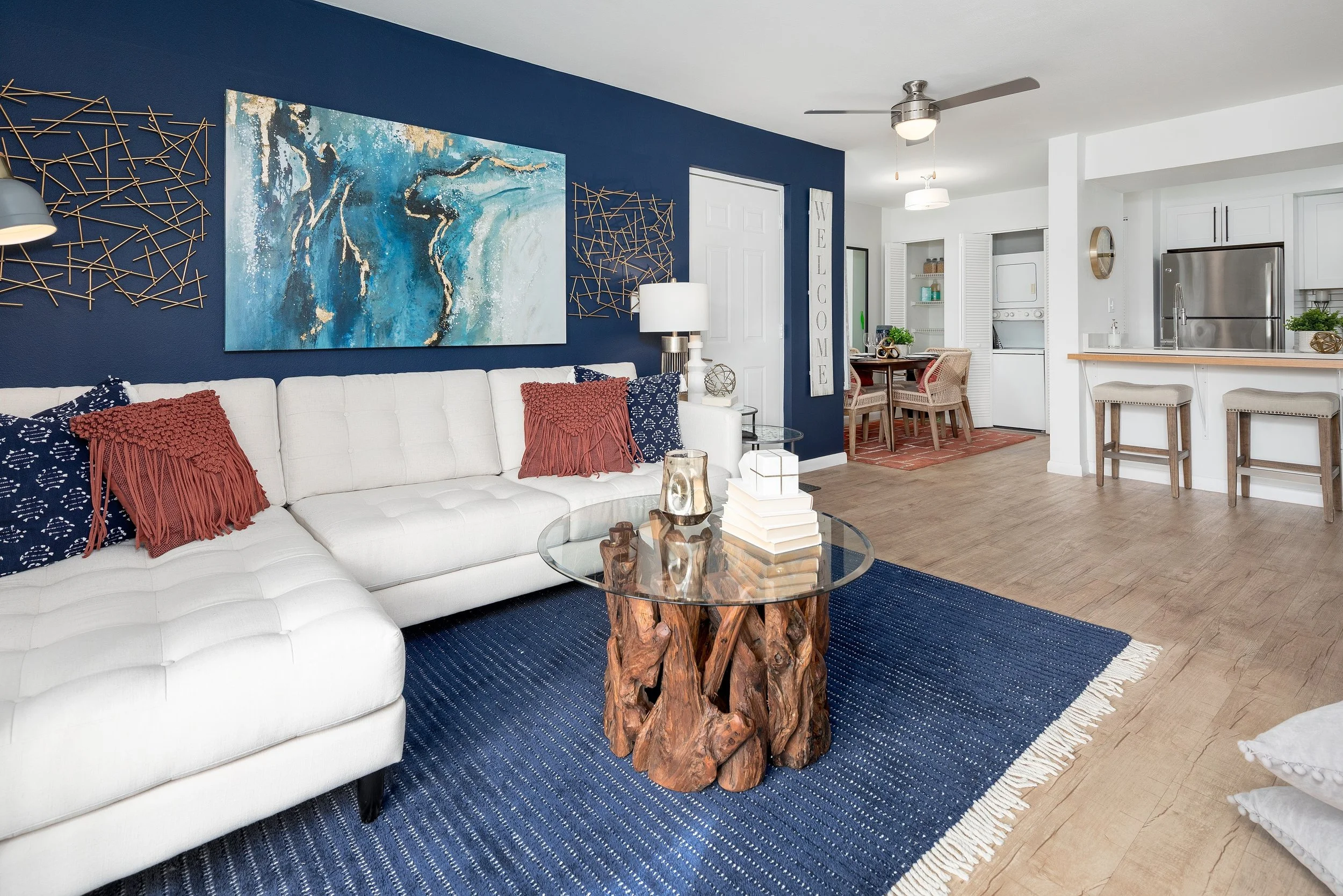 Living room with white sofa, blue walls, abstract artwork, wooden coffee table with glass top, and decorative pillows, open kitchen and dining area in background.