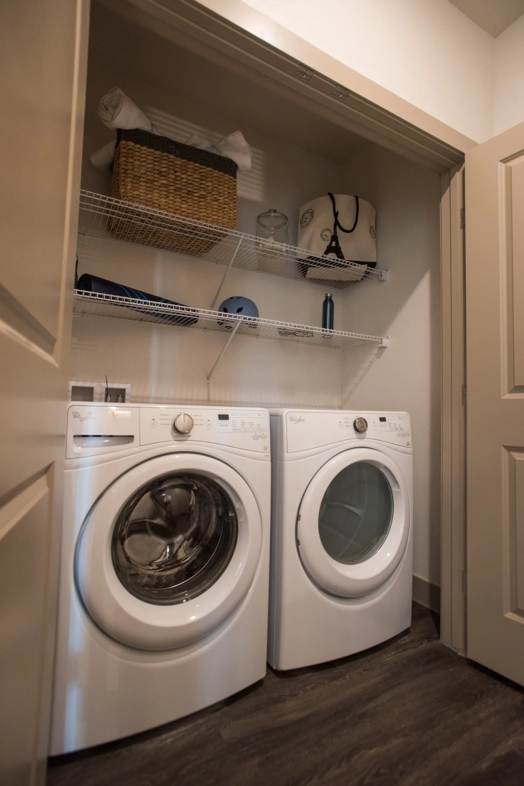 A laundry room with a front-loading washing machine and dryer, white in color, placed side by side. Above them, there are white wire shelves holding a woven basket with items inside, a glass jar, a purse with a Eiffel Tower print, a blue water bottle