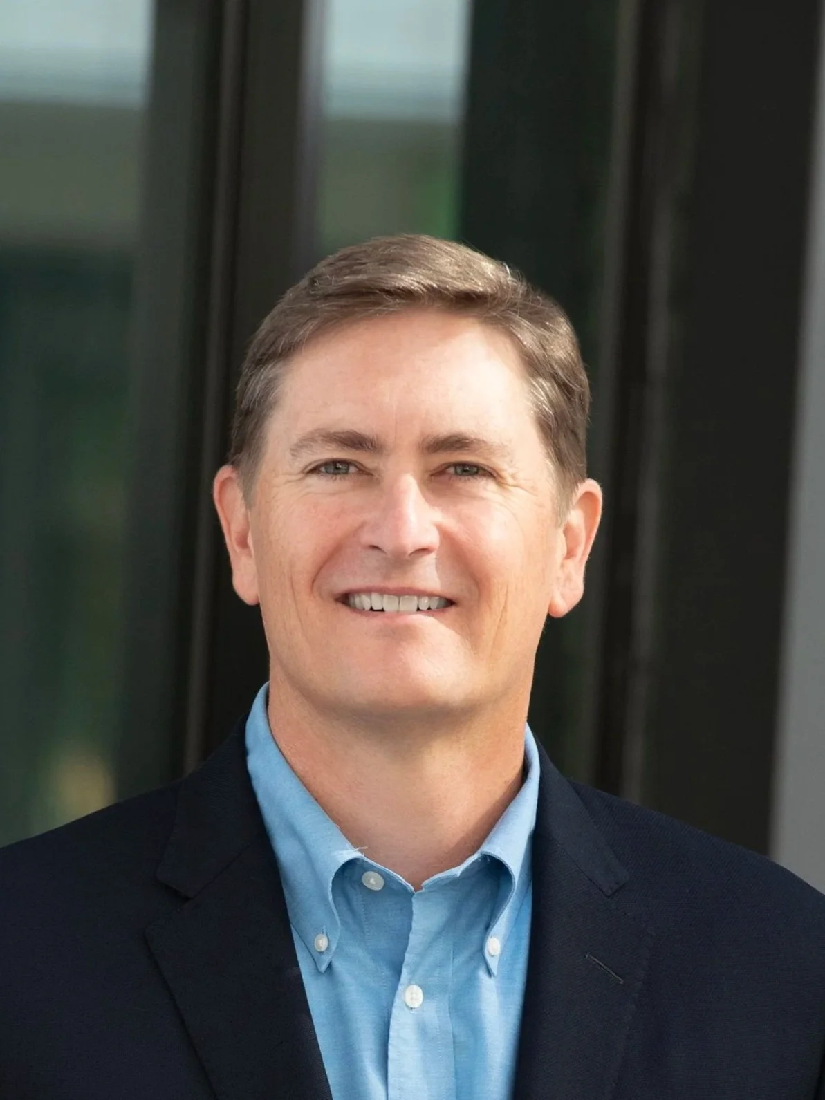 A portrait of a smiling man with short brown hair, wearing a blue dress shirt and a black blazer, standing outdoors with blurred background of glass windows.
