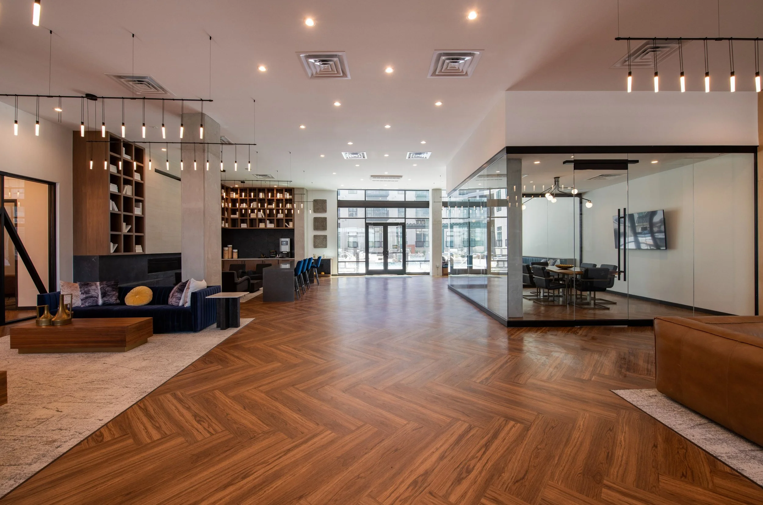 Spacious modern hotel lobby with wooden flooring, a sitting area with a velvet sofa and coffee table, and a glass-enclosed conference room on the right. Natural light streams from large front doors.