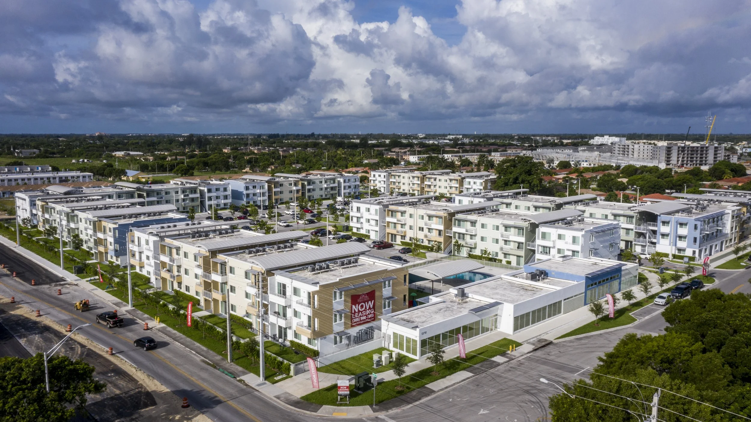 Aerial view of a modern apartment complex with white and beige buildings, a swimming pool, parking lots, and a road with cars and construction cones, under a partly cloudy sky.