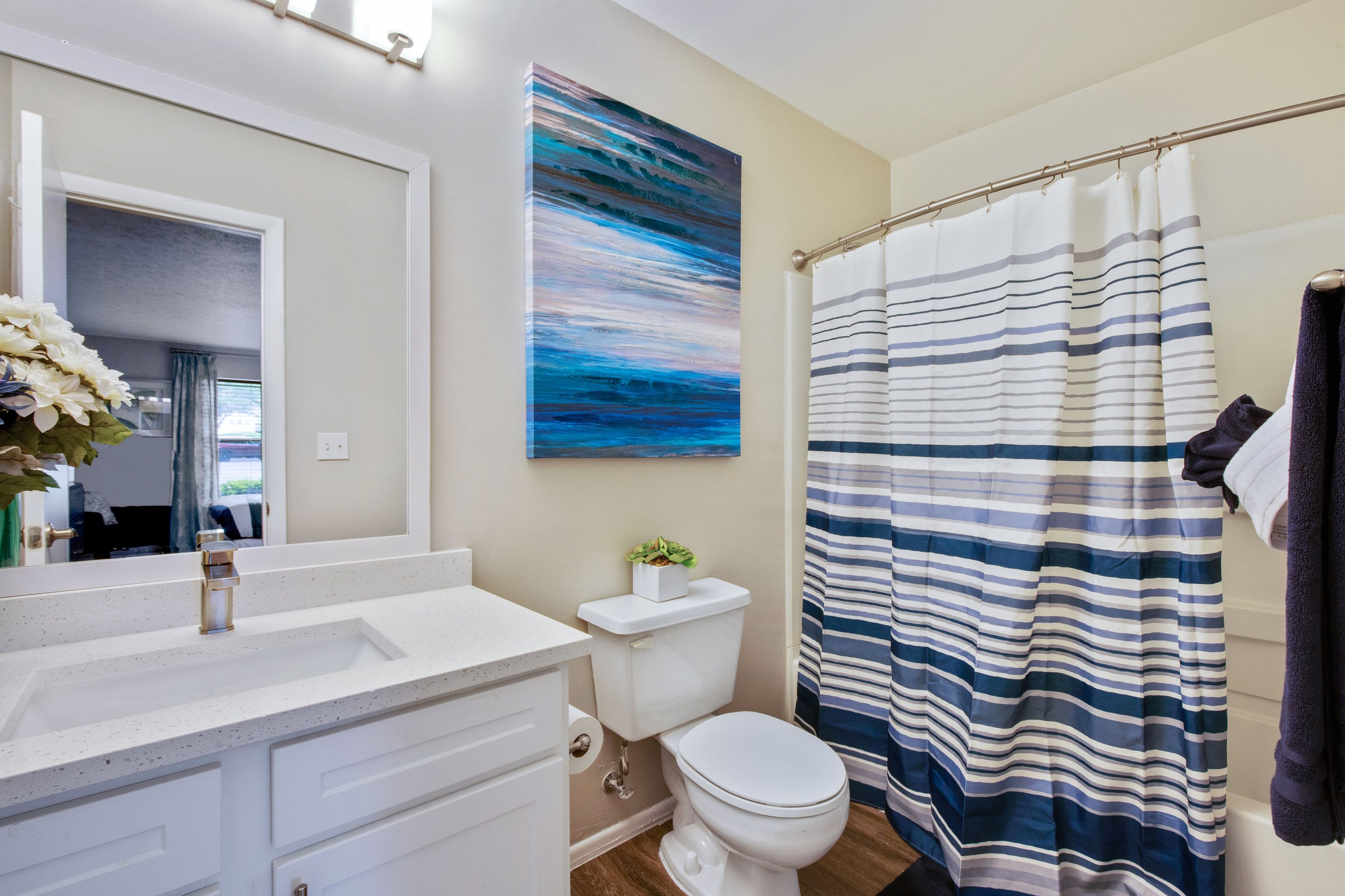 Bathroom with white vanity, large mirror, painting of ocean waves, toilet with plant on tank, striped shower curtain, towels on rack.