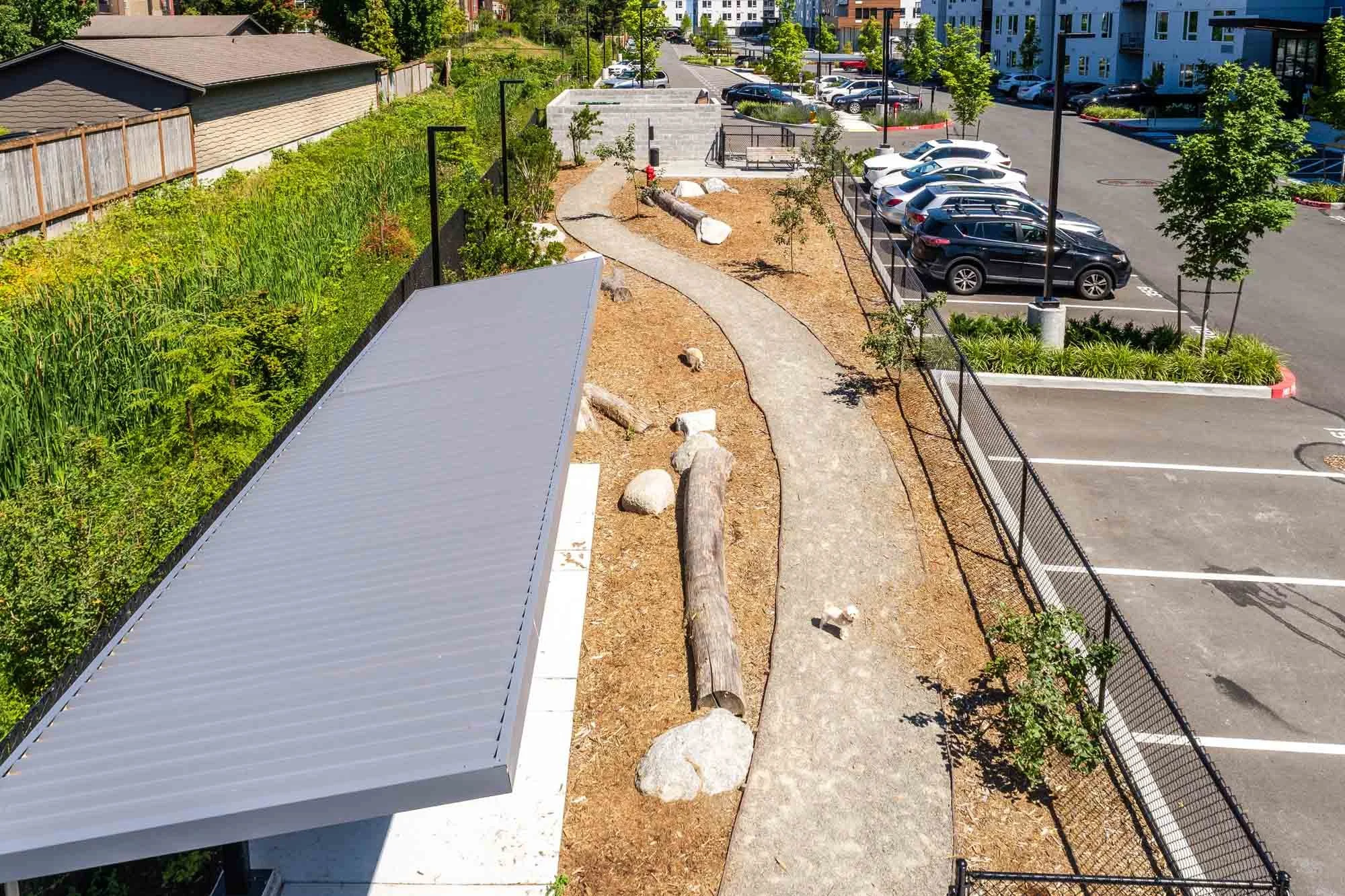 A winding concrete walkway runs through a landscaped area with trees, rocks, and logs beside a parking lot with cars, outdoor seating, and office or apartment buildings in the background.