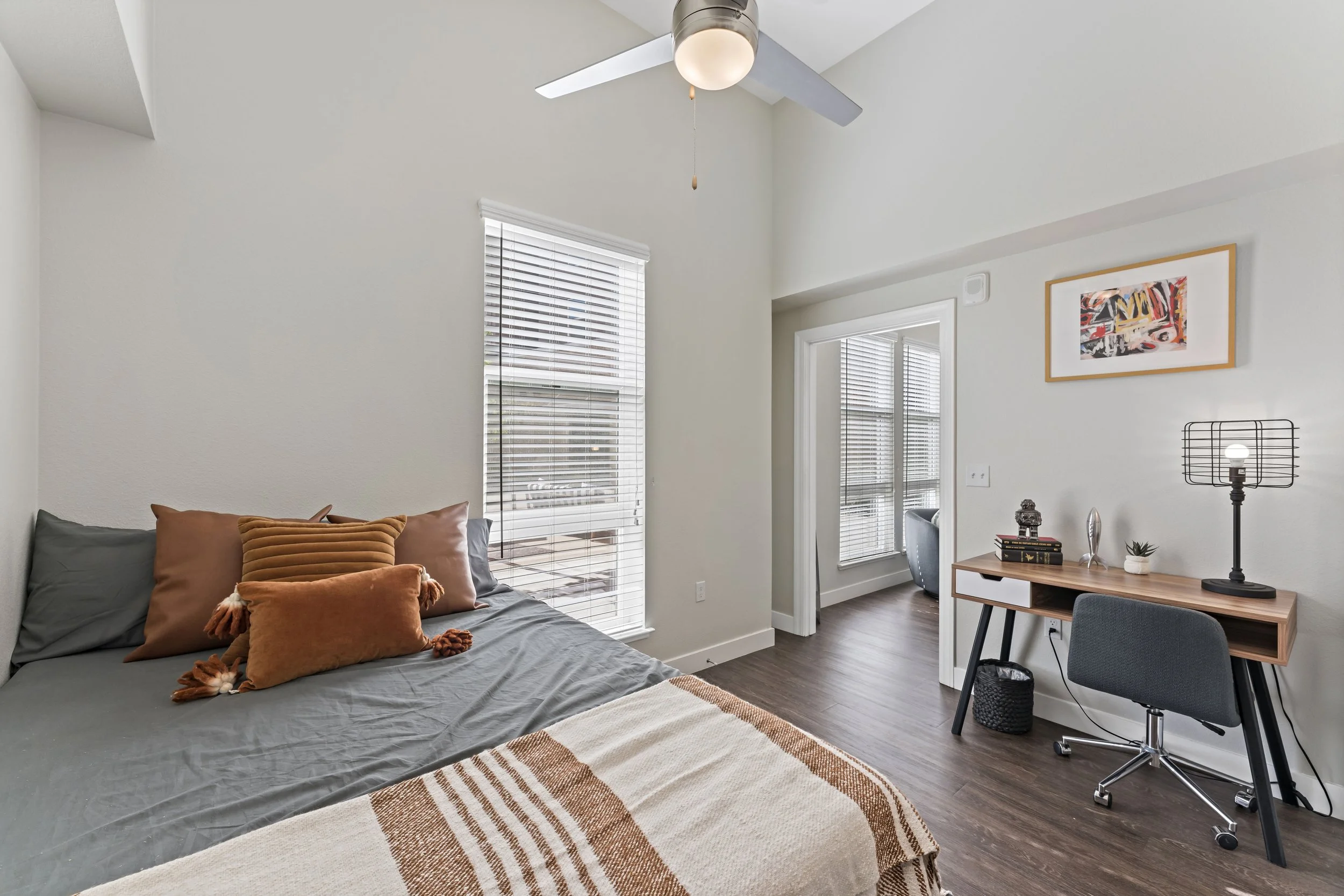 Bedroom with a bed, pillows, window blinds, a desk with a chair, table lamp, decorative items, and framed artwork on the wall.
